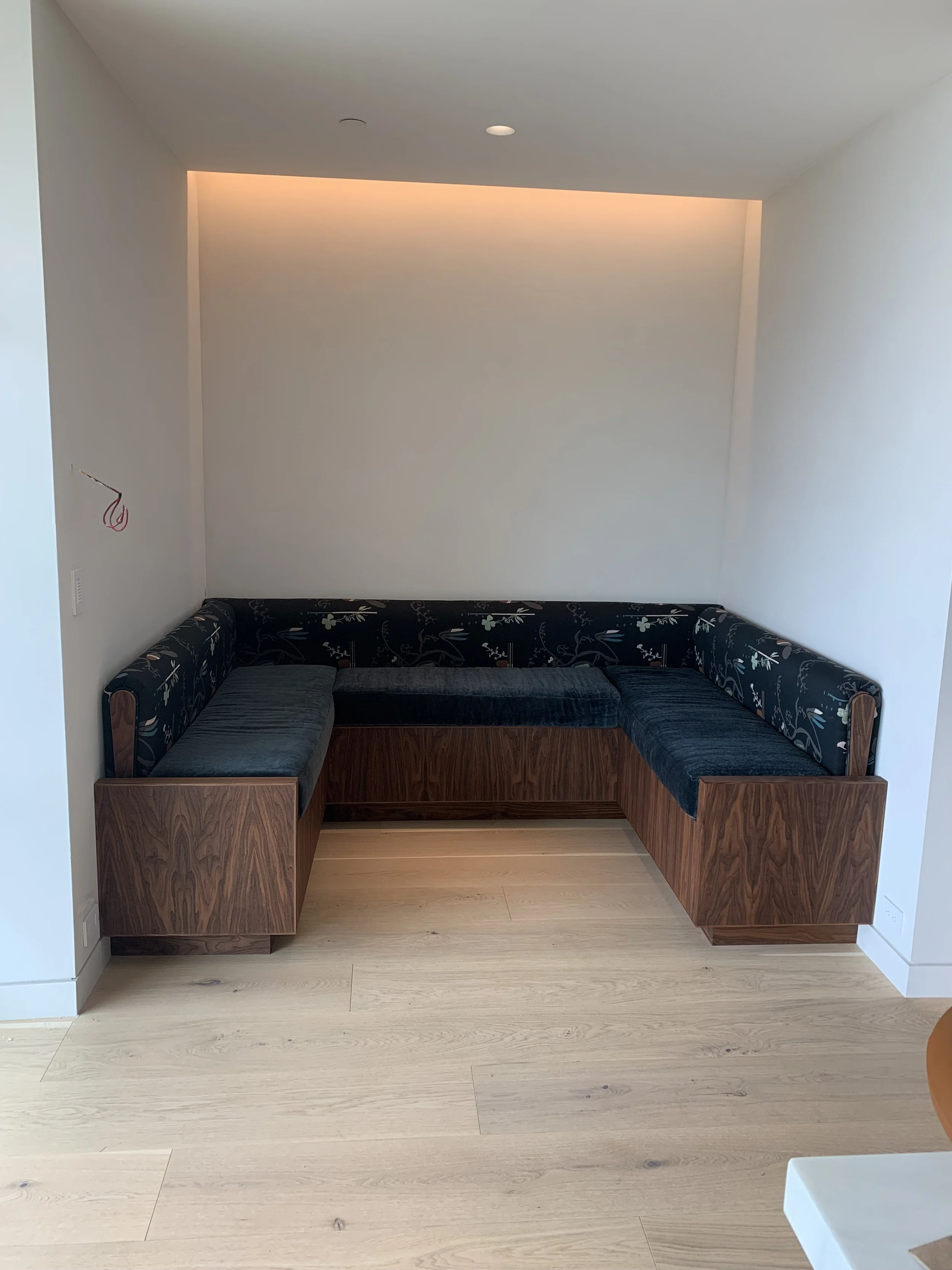 Corner seating area with a wooden built-in bench and cushioned seats with dark floral upholstery in a minimalist room with light wood flooring and white walls.
