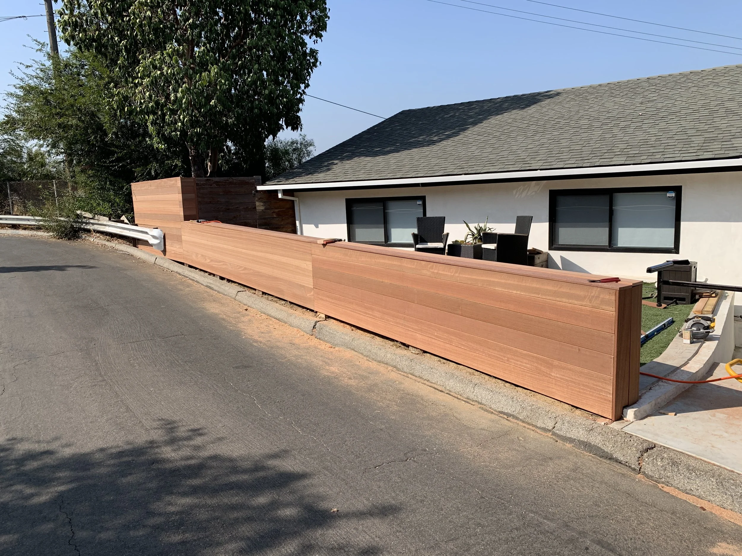 A house with two large windows and a gray roof, with an outdoor patio area featuring two black chairs, a small table with a plant, and construction tools including a saw and level, and a long wooden fence being built along the sidewalk.