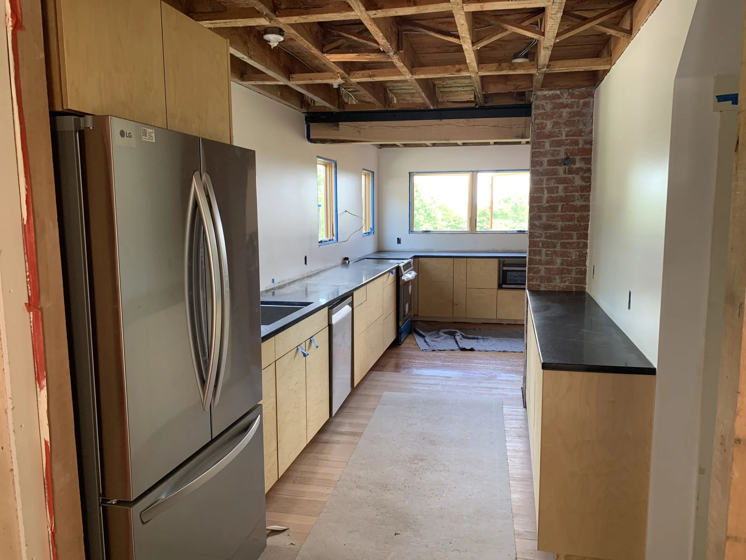 Kitchen under construction with wooden cabinets, stainless steel refrigerator, black countertops, and brick wall segment, with two windows letting in natural light.