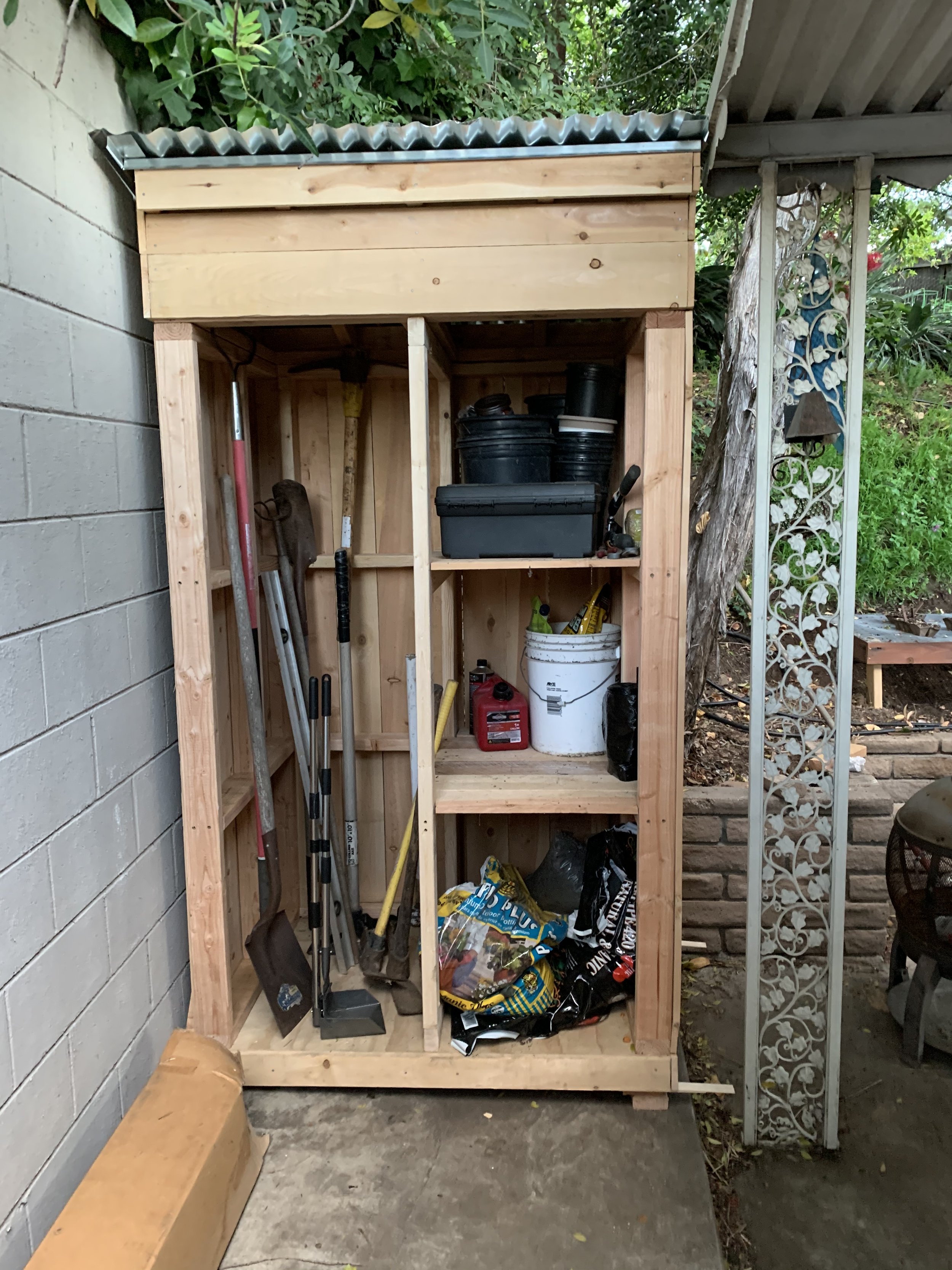 A wooden outdoor storage shed filled with gardening tools, black containers, a white bucket, and various supplies, situated near a brick and concrete patio area.