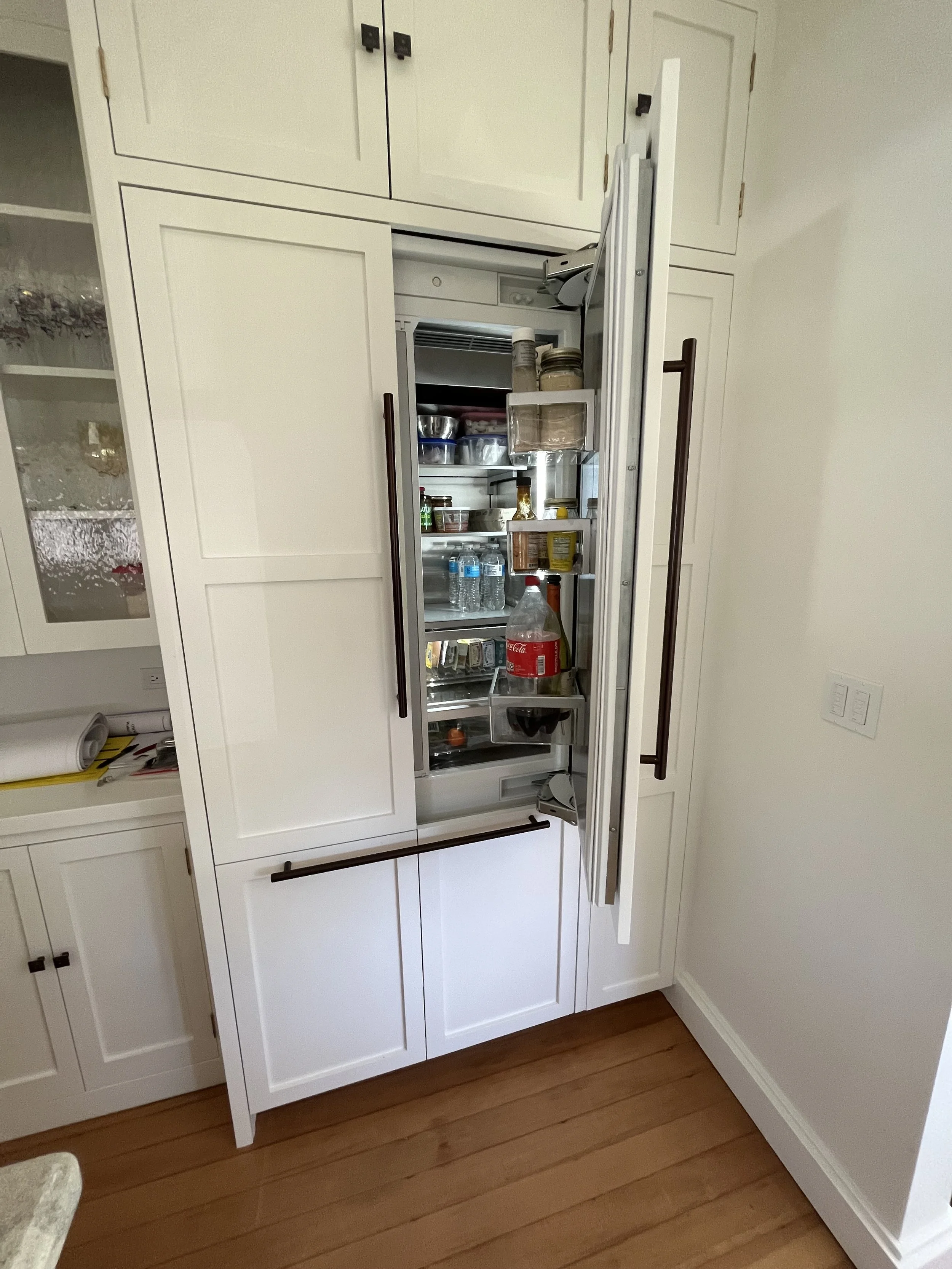 Open refrigerator in a kitchen with items like water bottles, condiments, and beverages inside, integrated into white cabinetry and a hardwood floor.
