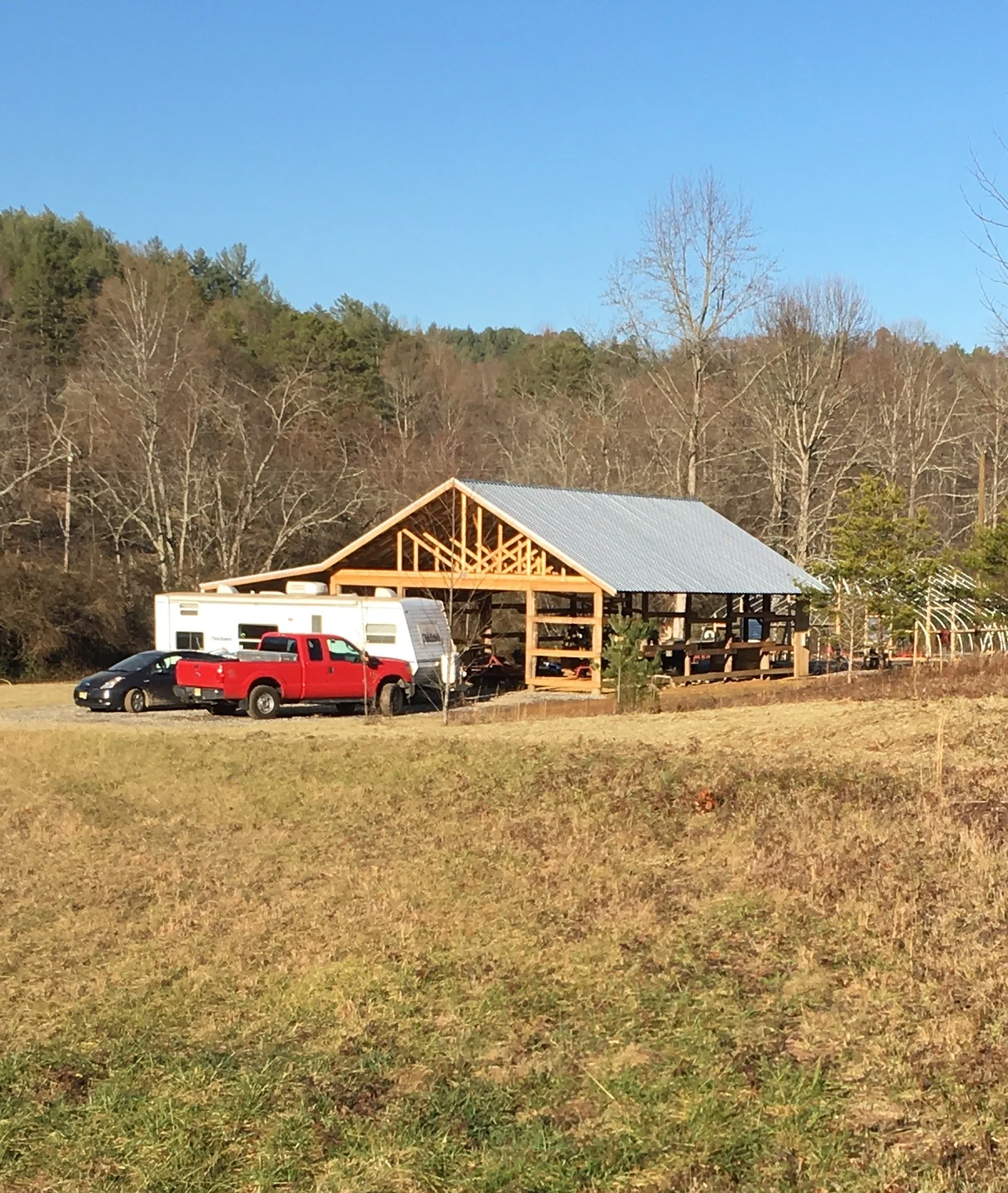 A partially constructed wooden building with a metal roof, surrounded by a landscaped yard with a red truck, a white trailer, and a black car parked nearby, set against a background of leafless trees and a blue sky.