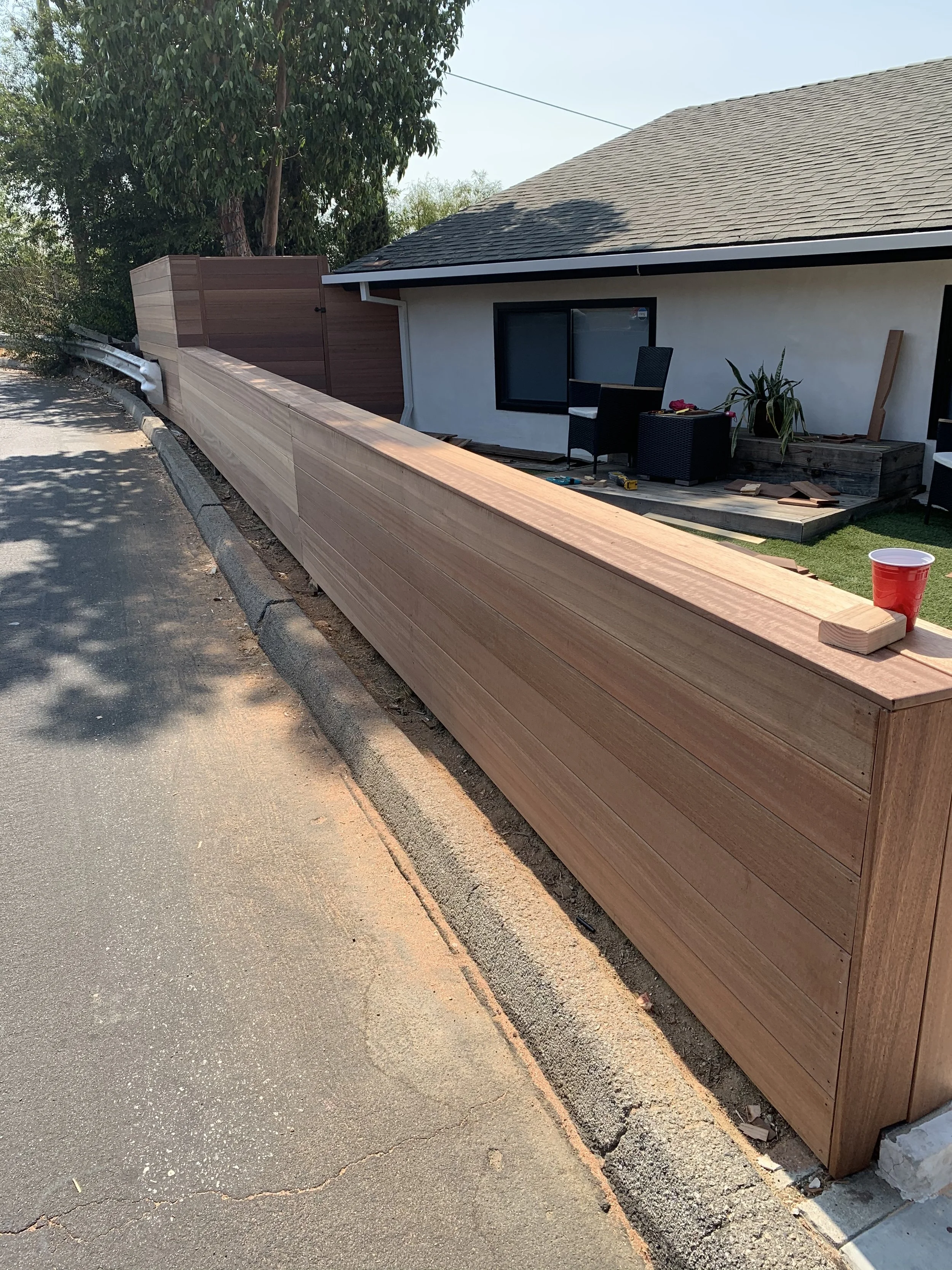 A long wooden fence under construction next to a house with patio furniture and a potted plant, on a sidewalk with trees in the background.