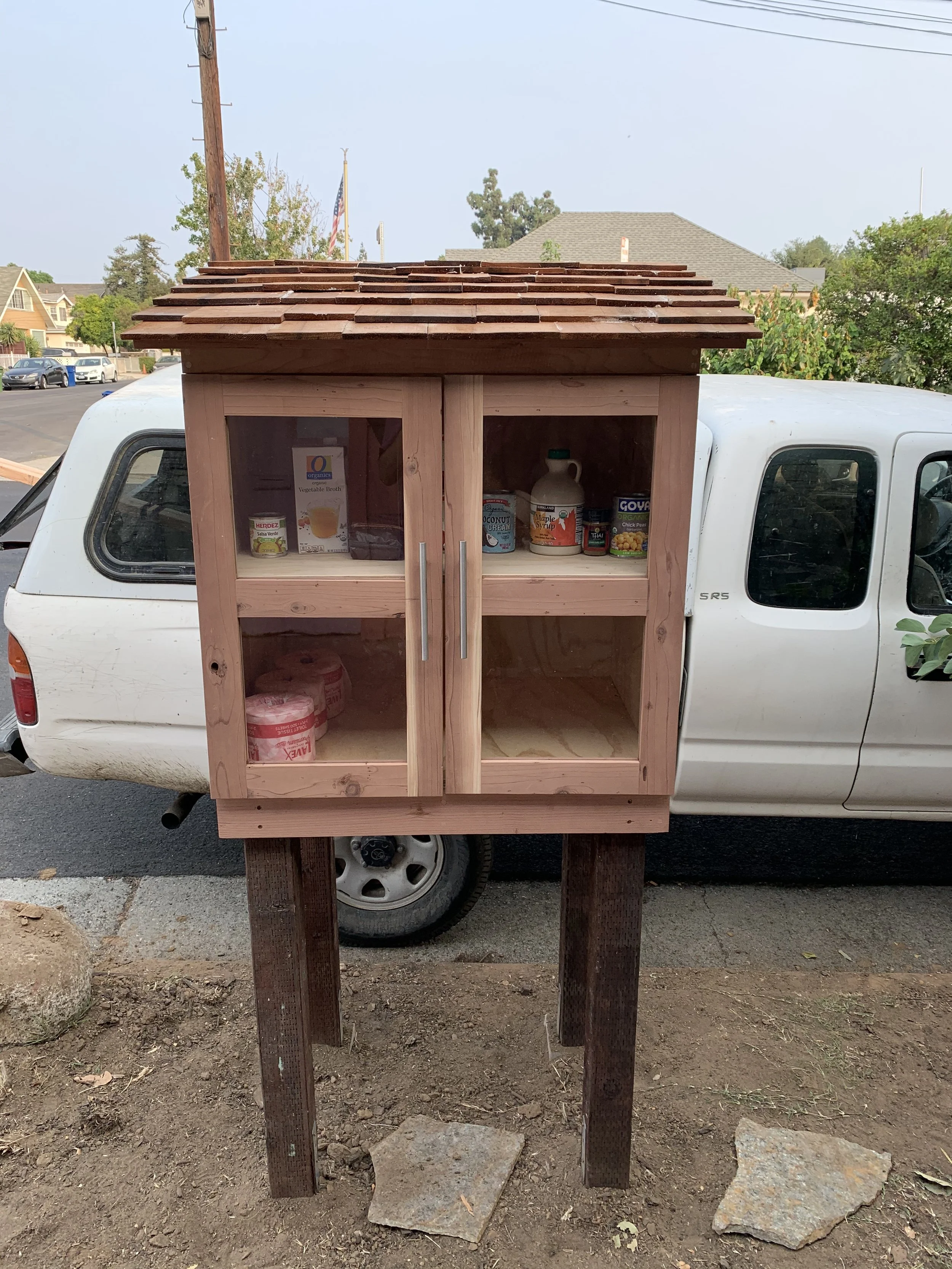 A small wooden food pantry mounted on a white truck, with shelves containing canned goods, bottled syrups, and food items.