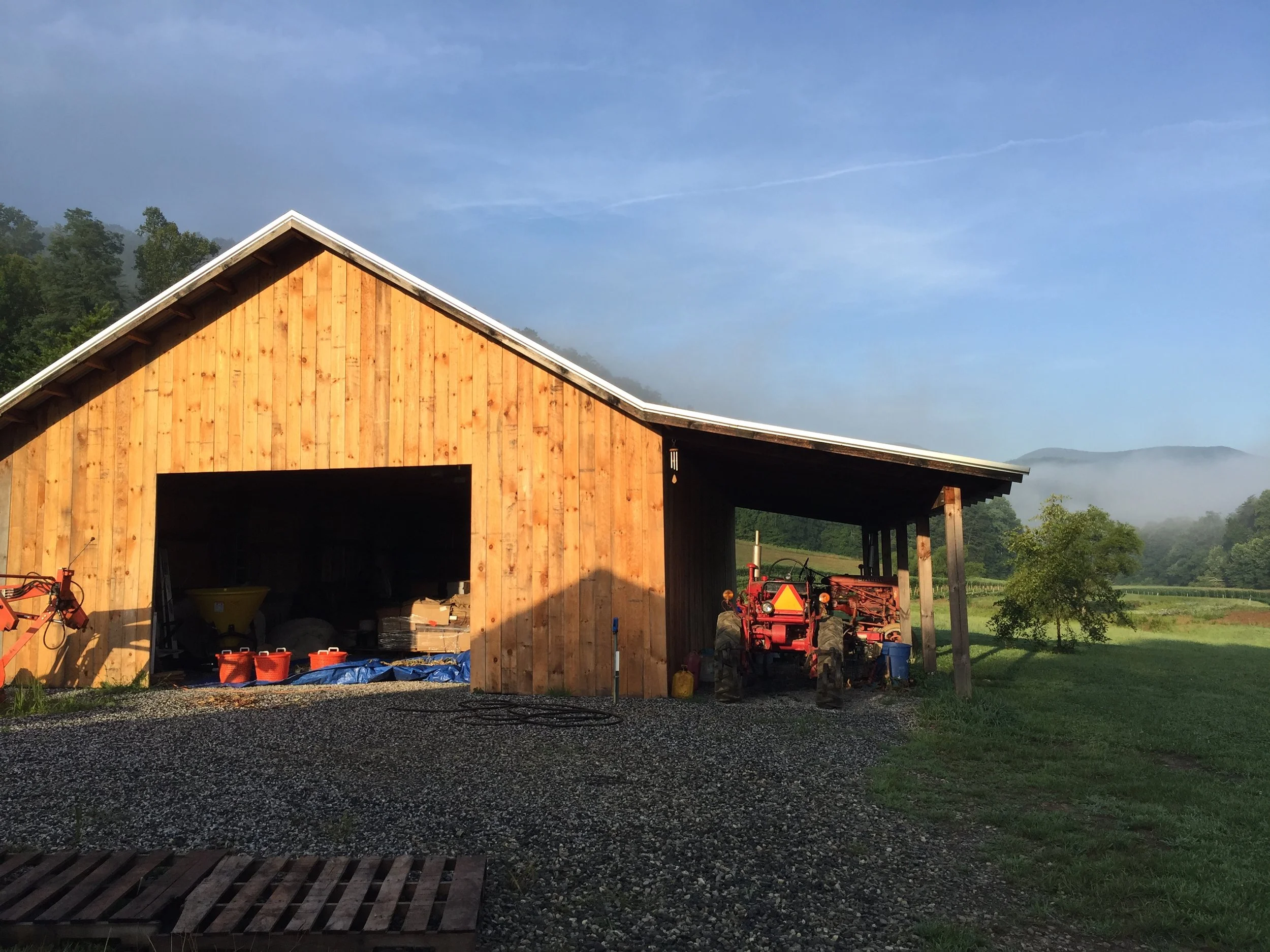 A wooden barn with an open front, a red tractor parked underneath its roof, and surrounded by green fields with trees and distant hills.