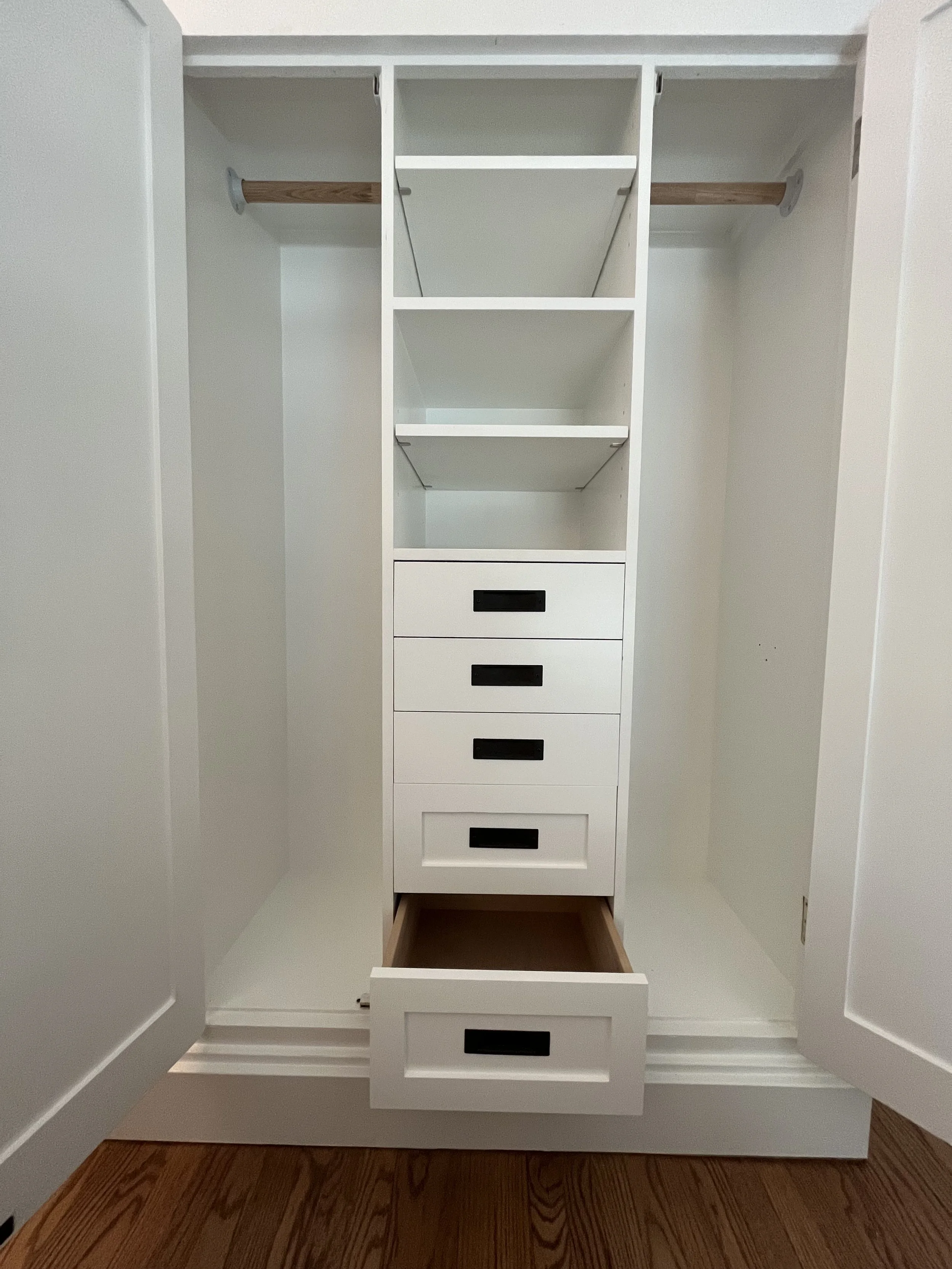 Empty white closet with five drawers, open bottom drawer, two shelves, and hanging rods on each side, on a hardwood floor.