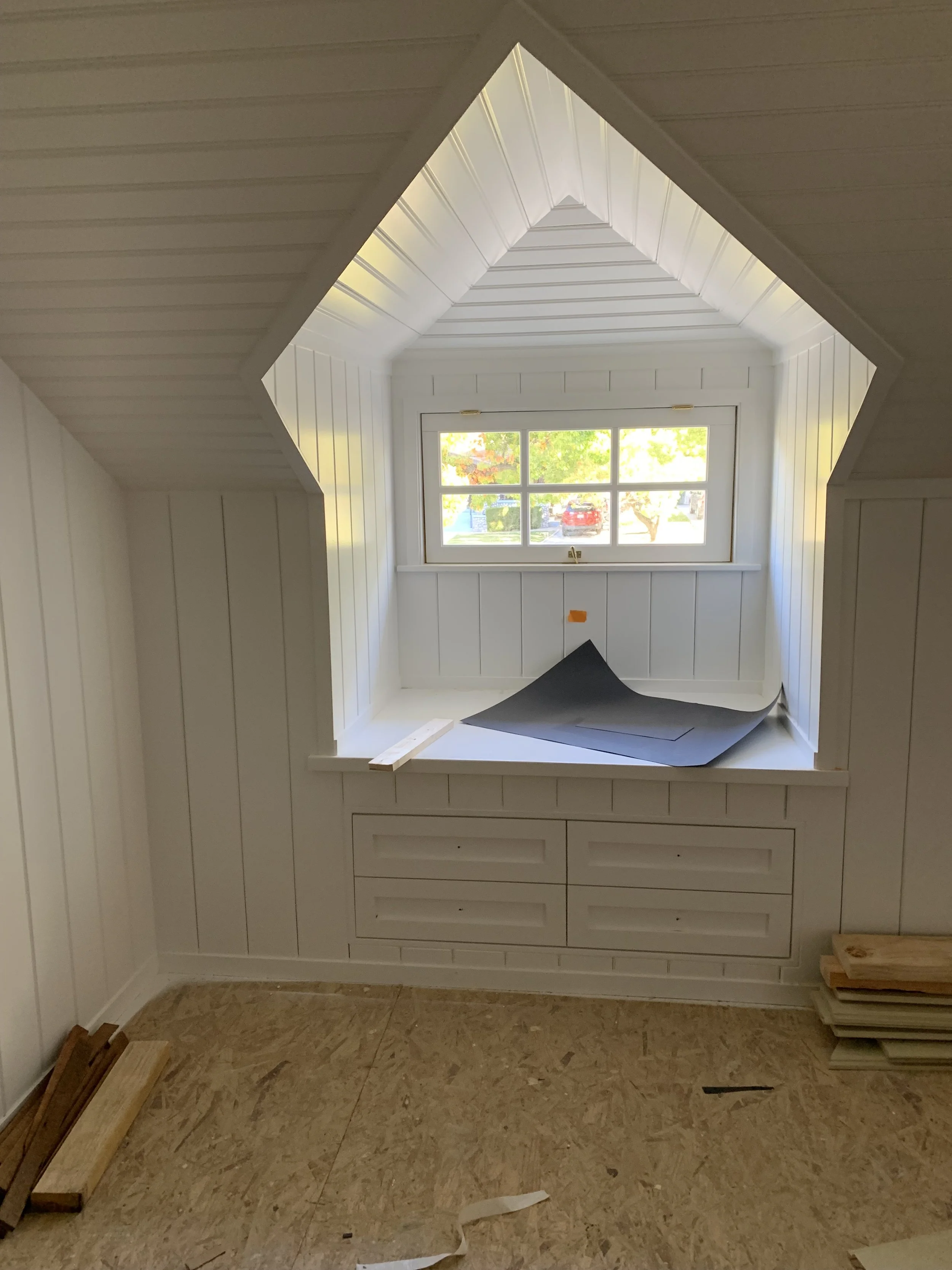 A small attic room with a painted white wooden wall and a window showcasing a neighborhood with trees and cars. Construction materials and black foam sheets are scattered around.