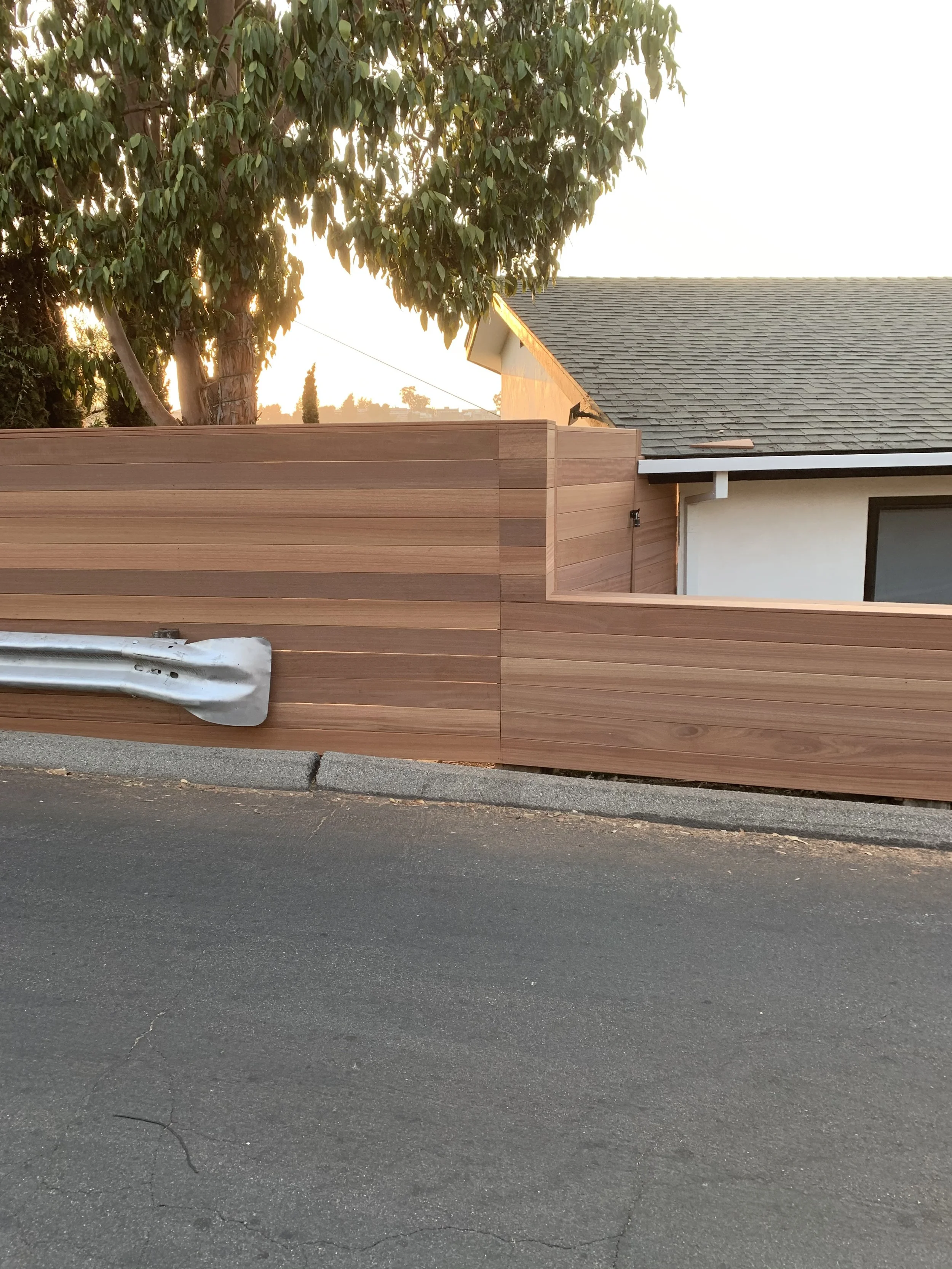A wooden fence made of horizontal planks runs along the sidewalk in front of a house with a gray shingle roof and white walls, with a large tree behind the fence and sunlight filtering through the leaves.