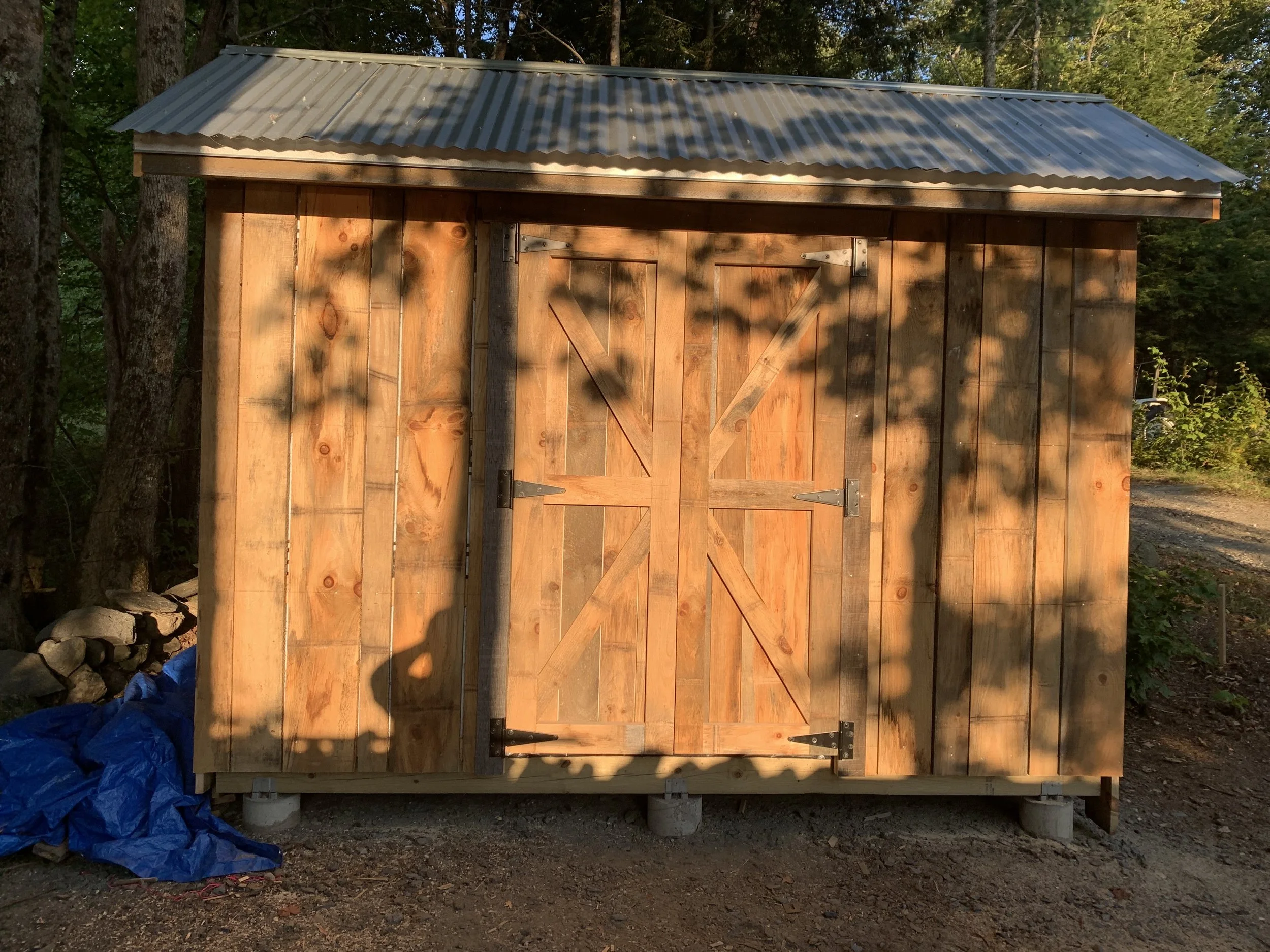 A small wooden shed with a metal roof, located outdoors on a dirt ground surrounded by trees and bushes, casting shadows onto its front.