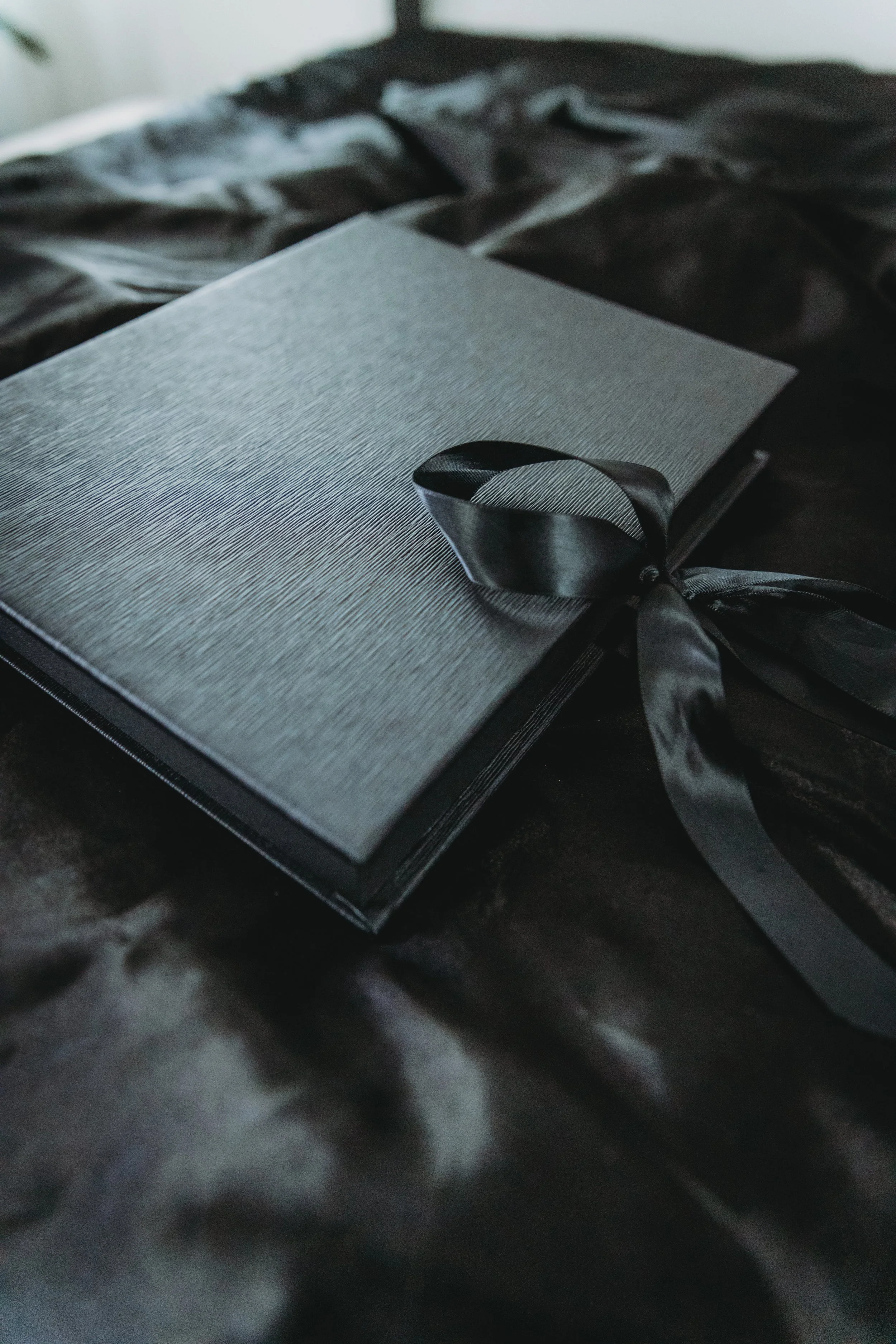 A closed black gift box with a textured surface and a black satin ribbon tied in a bow, placed on a black satin fabric surface.