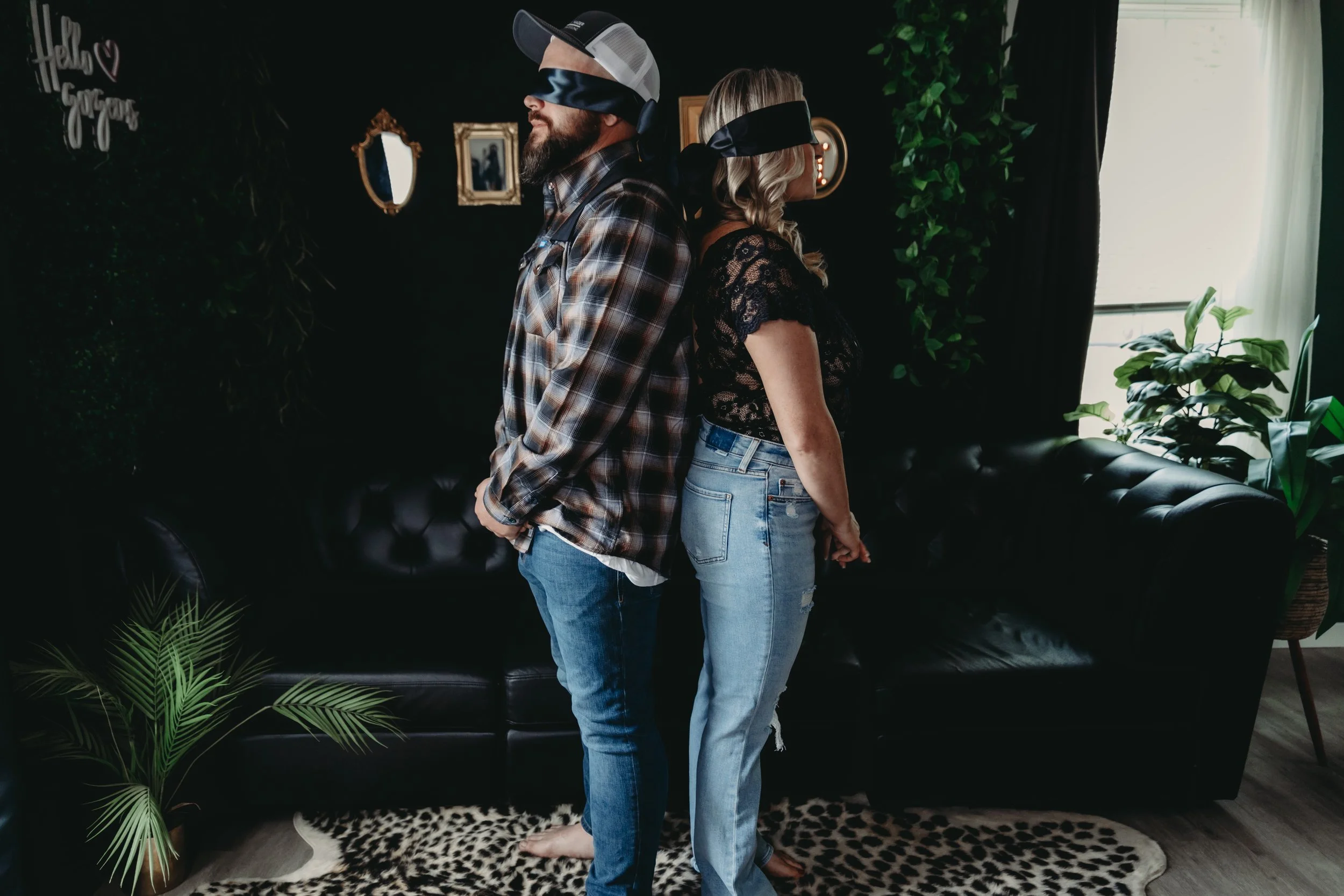 A man and woman stand back-to-back in a living room, both blindfolded with black fabric. The man wears a plaid shirt and jeans, while the woman wears a lace top and jeans. They are barefoot, and there are various plants and framed pictures on the dark wall behind them.