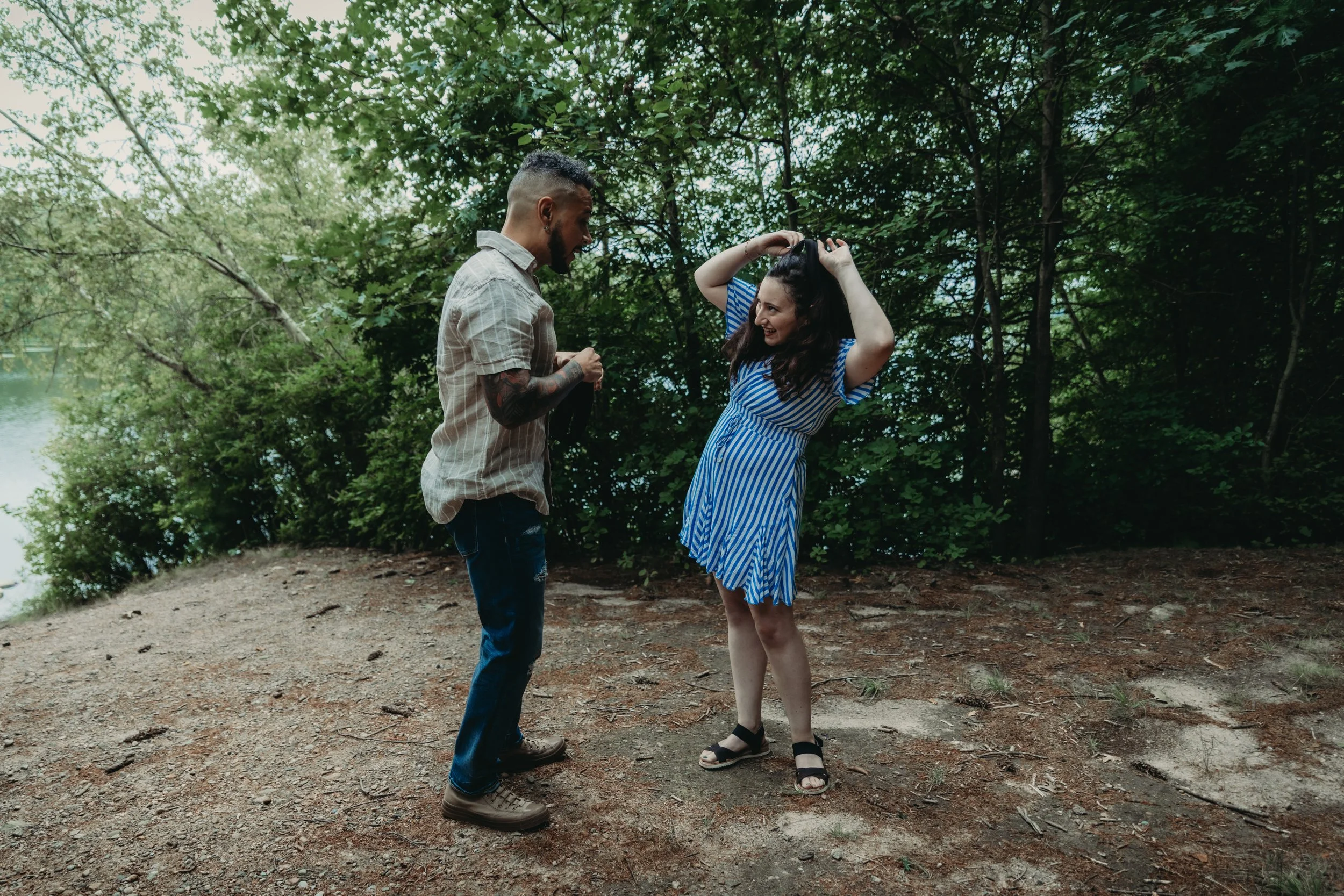 A man and woman standing outdoors on a dirt path surrounded by green trees, engaging in conversation. The woman is adjusting her hair, and the man is holding sunglasses.