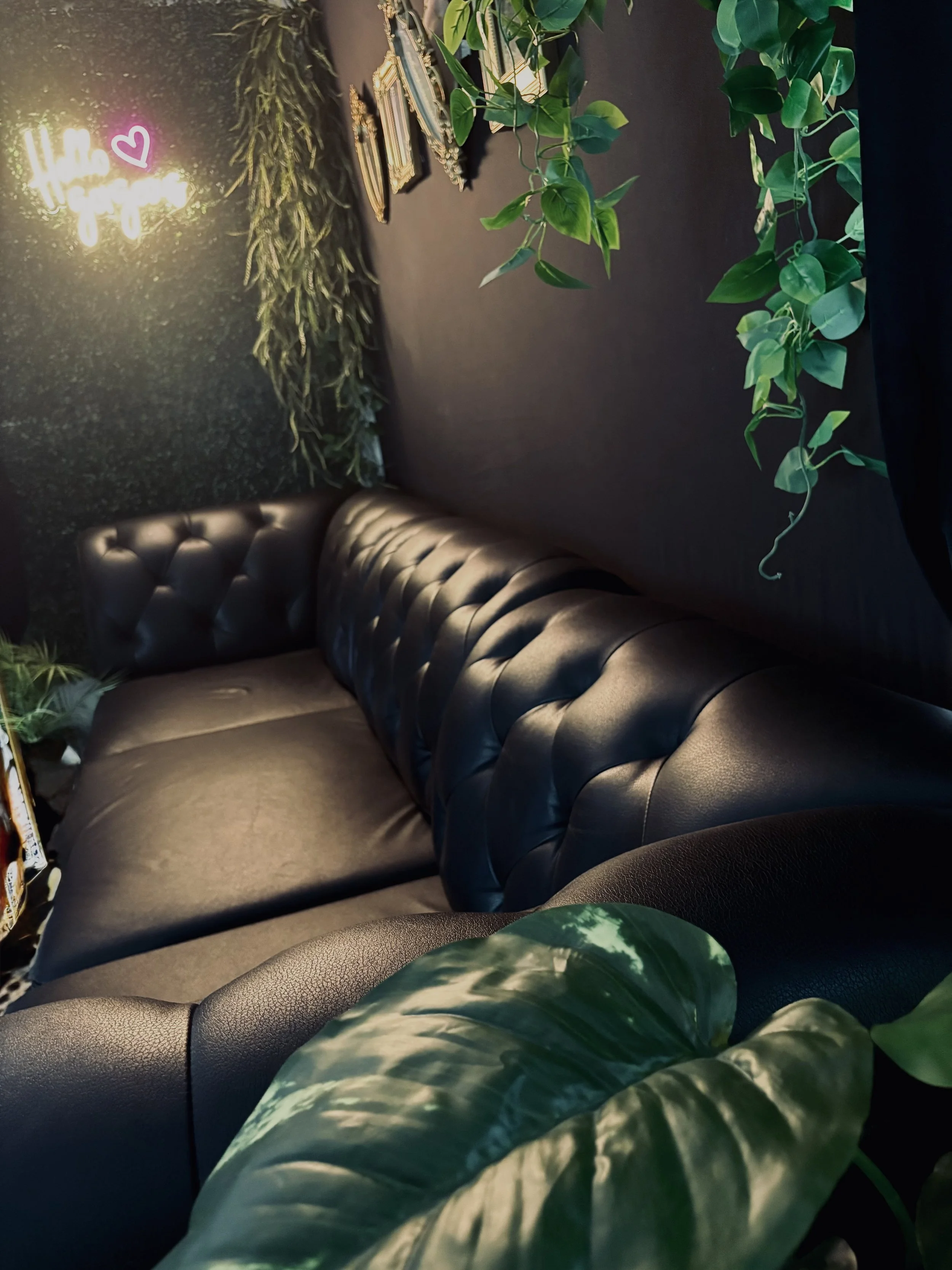 A black leather tufted sofa against a dark wall decorated with hanging green plants. On the wall, there's a neon sign that says 'Hello Gorgeous' with a pink heart. Several picture frames are also hanging on the wall.