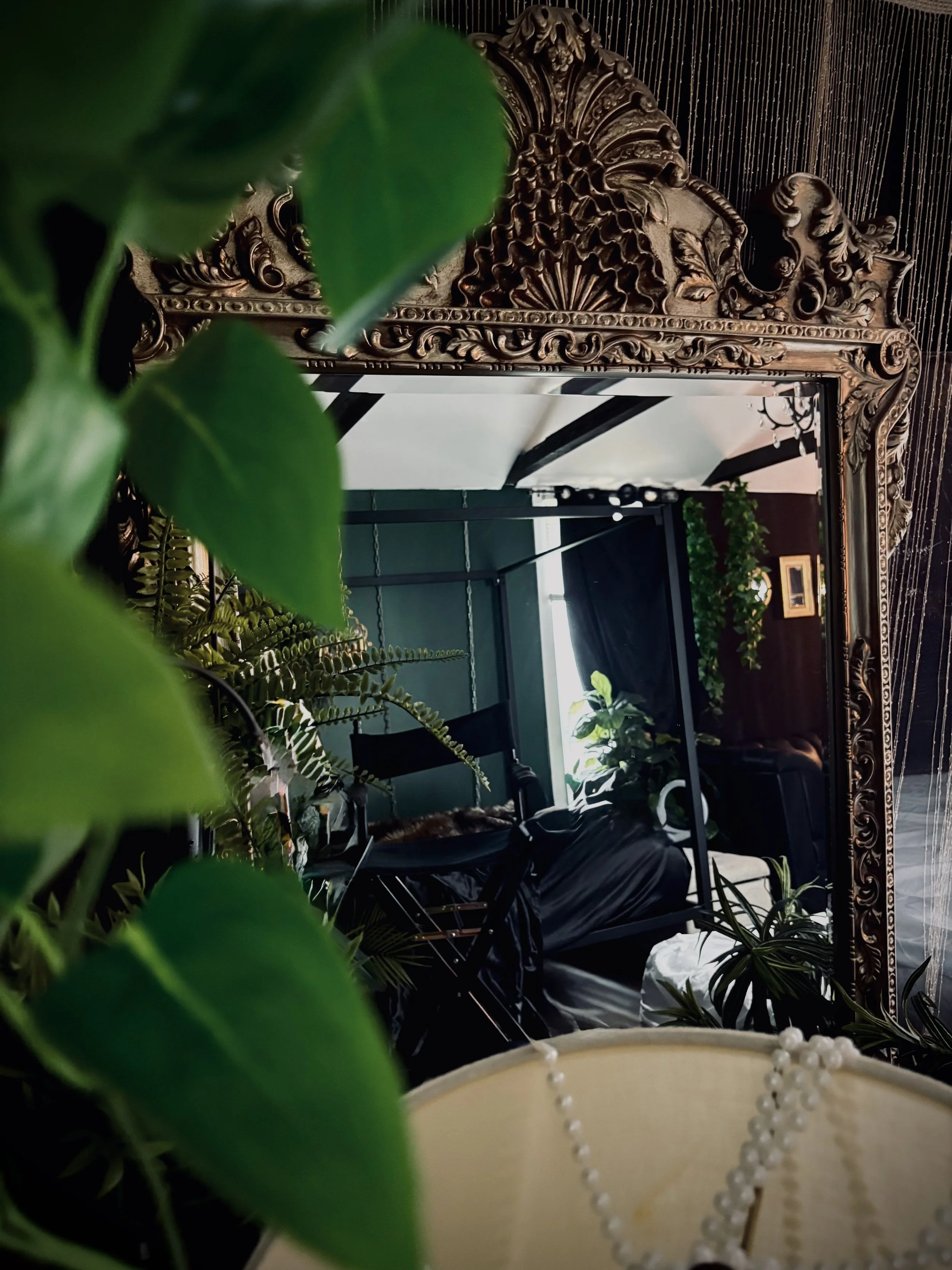 A mirror with an ornate, carved wooden frame reflects a room with green/pine-colored walls, plants, black furniture, and a bright window.
