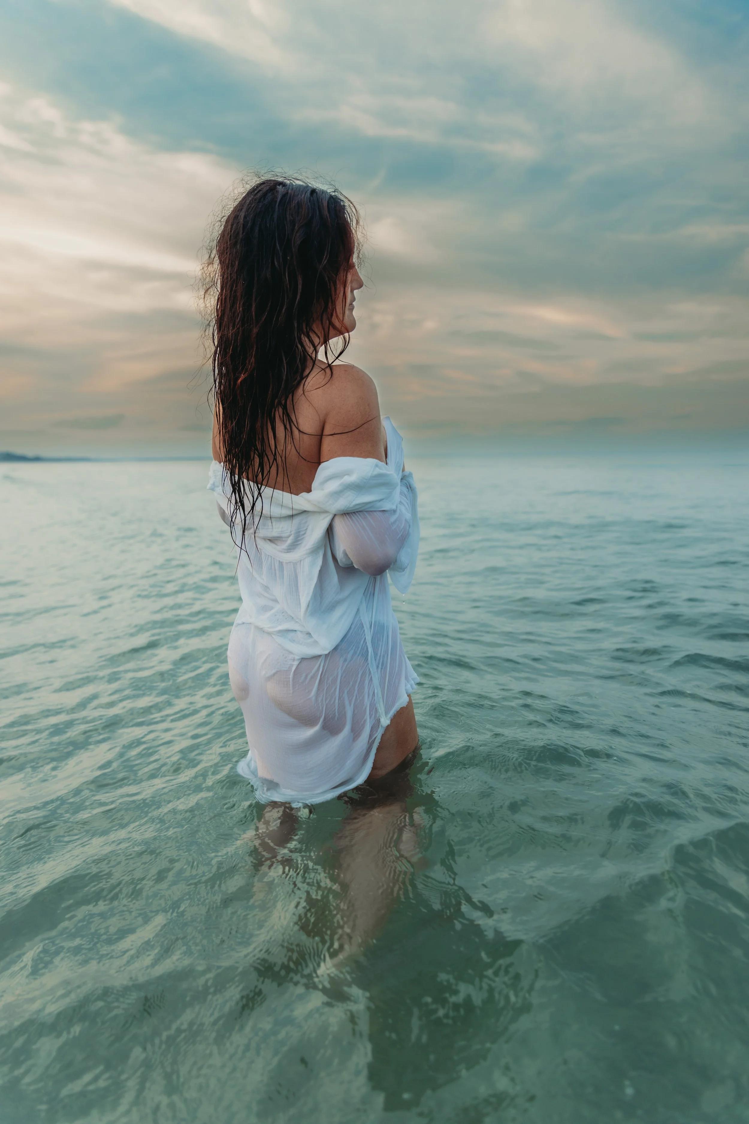 Woman in white, wet dress standing in shallow ocean water with cloudy sky in the background.