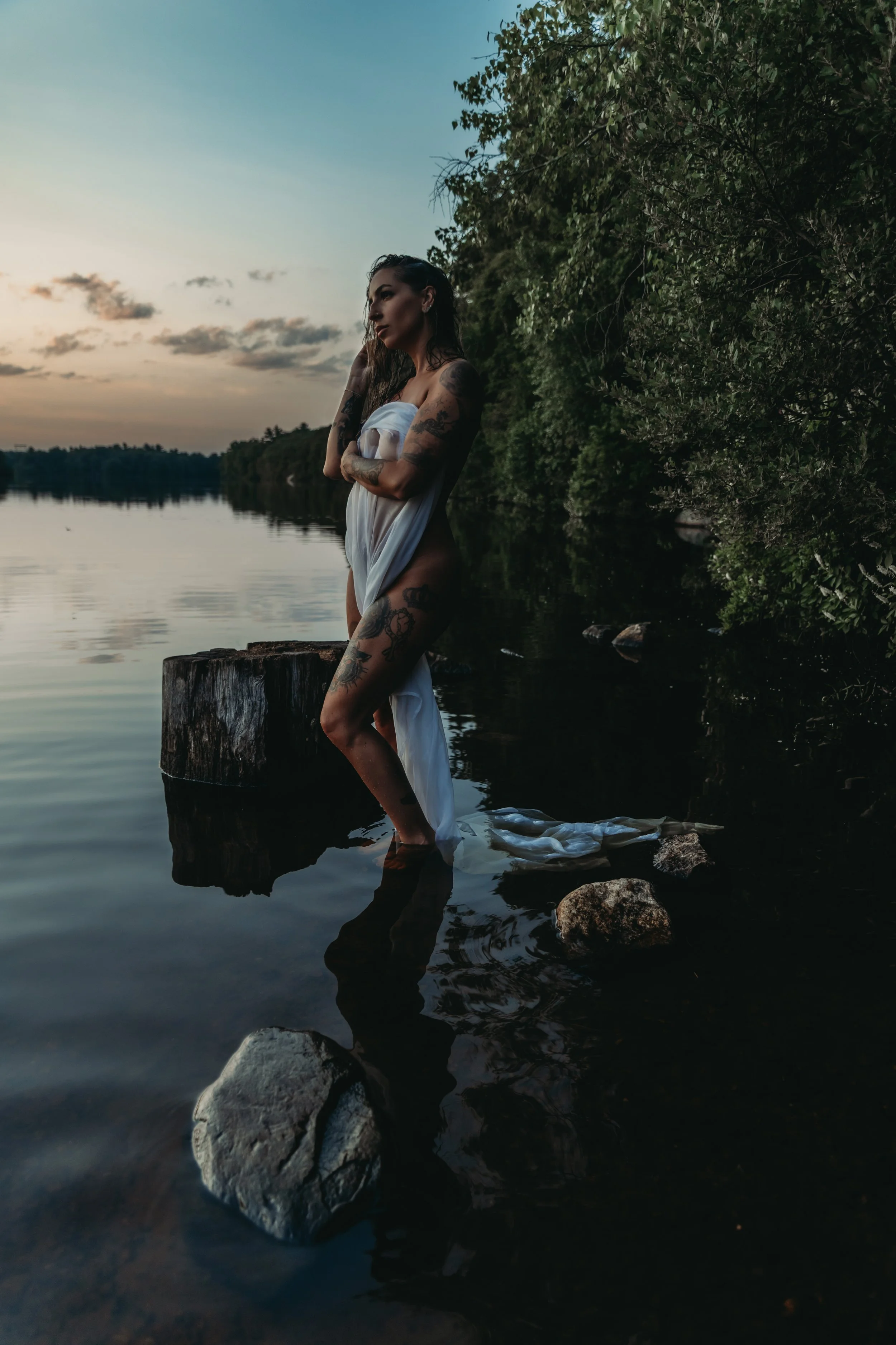 A woman with tattoos standing in the water at sunset near a forested shoreline, wrapped in a white cloth.