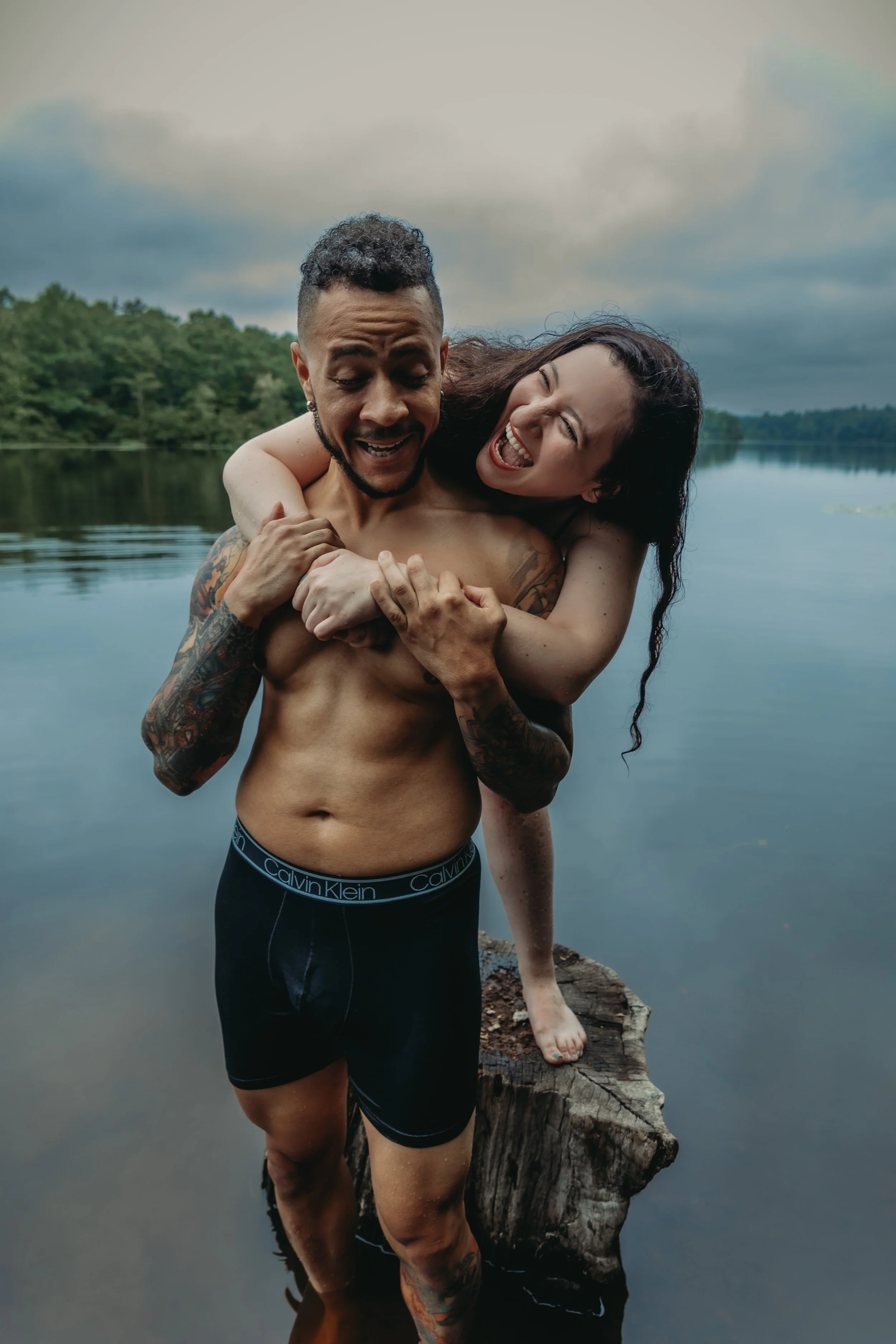A man and woman near a lake, with the man carrying the woman on his back, both smiling and laughing, on a cloudy day.