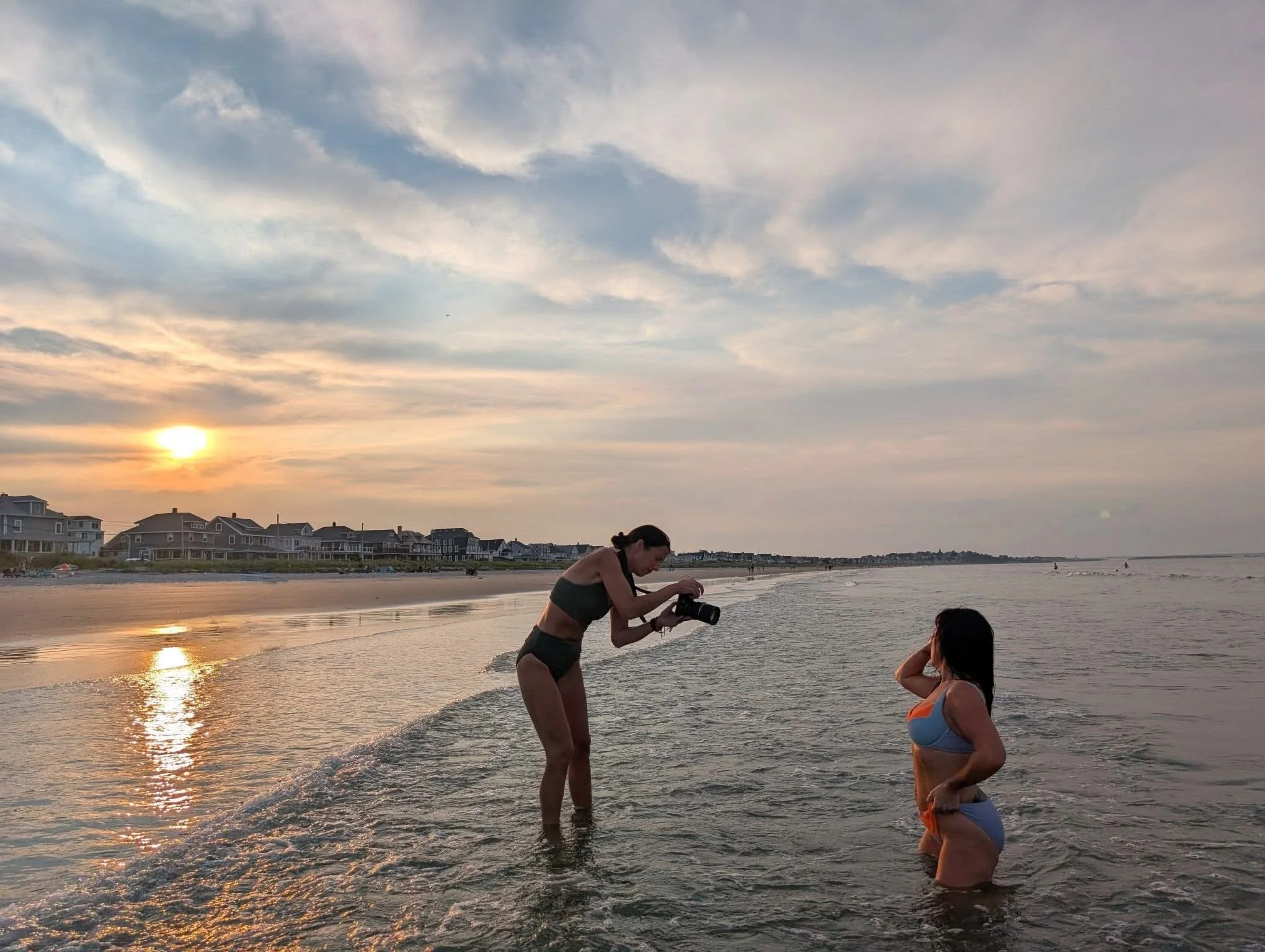 A woman in a swimsuit takes a photo of another woman in a swimsuit in the water at the beach during sunset.
