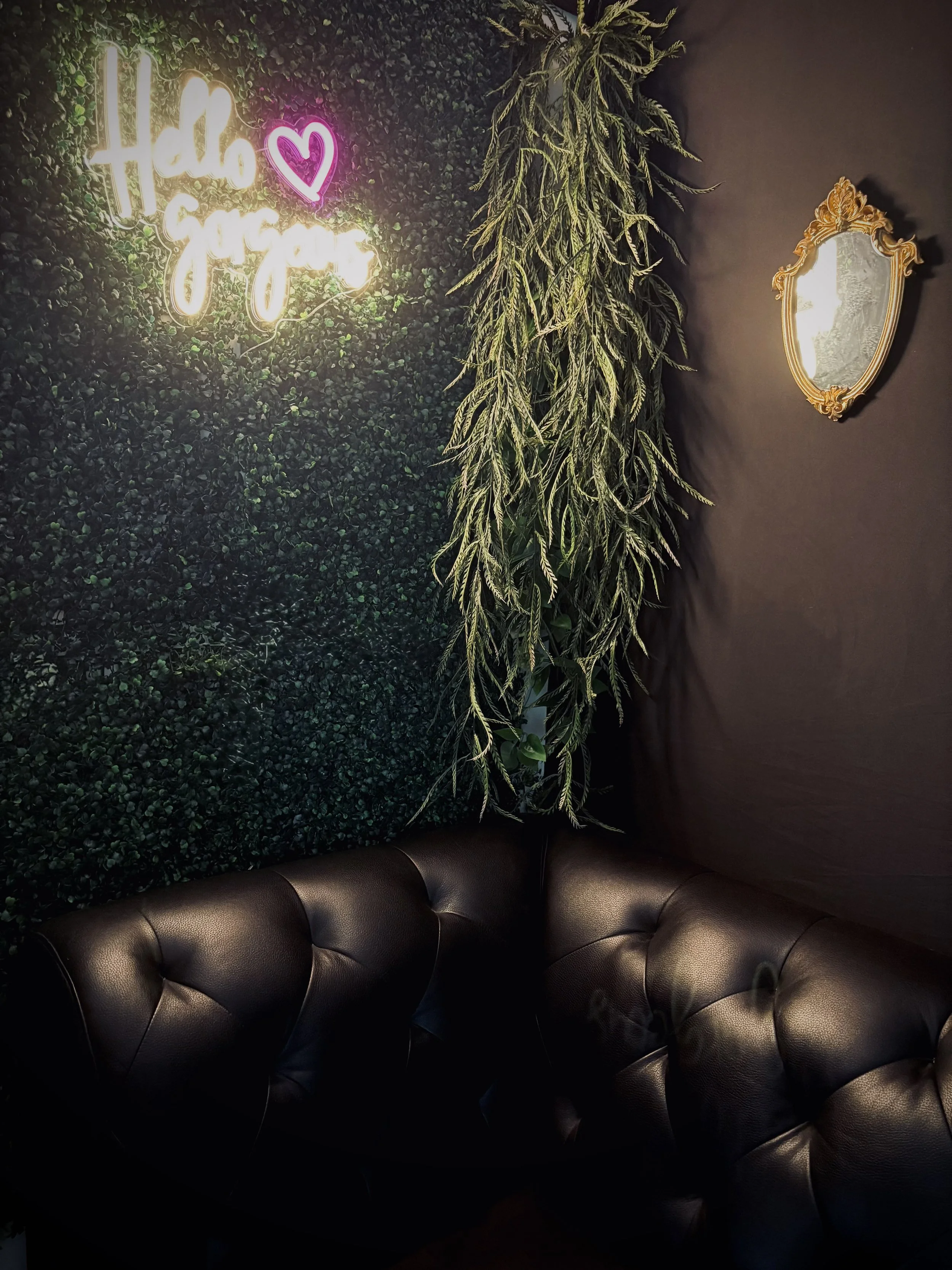 A corner of a room with a green leafy wall and a dark wall. A neon sign that says "Hello Gorgeous" with a pink heart. Two black tufted leather chairs. A large hanging plant. An oval mirror with a gold ornate frame.