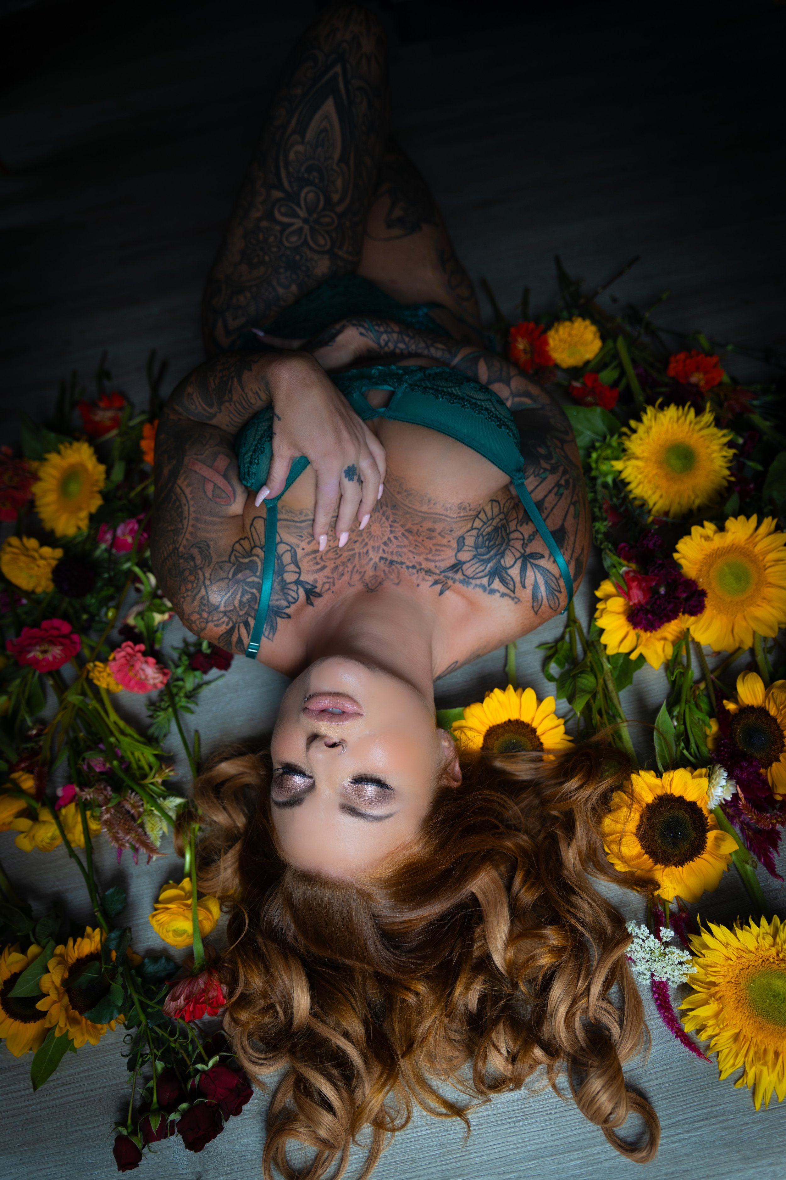 A woman with red hair, tattoos, and makeup lying on a wooden floor surrounded by various colorful flowers, including sunflowers.