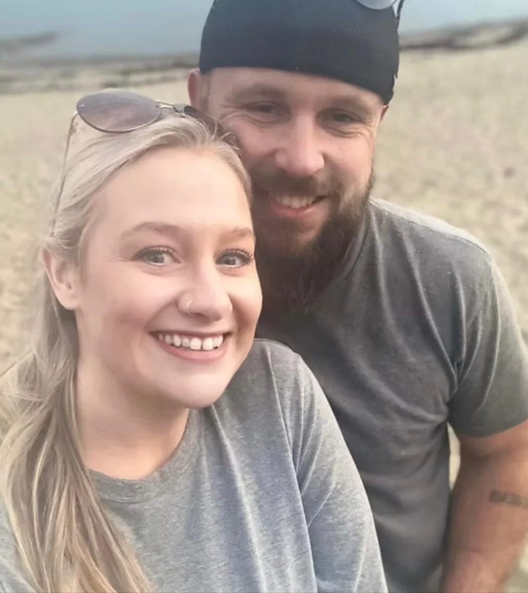 A smiling blonde woman with sunglasses on her head and a bearded man in a black cap, both wearing gray t-shirts, taking a selfie outdoors on a beach.