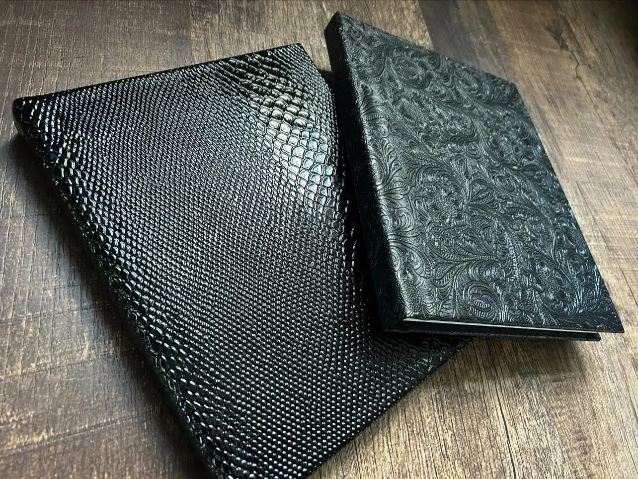 Two black purses with textured patterns, one resembling snake skin and the other with floral or paisley designs, lying on a wooden surface.
