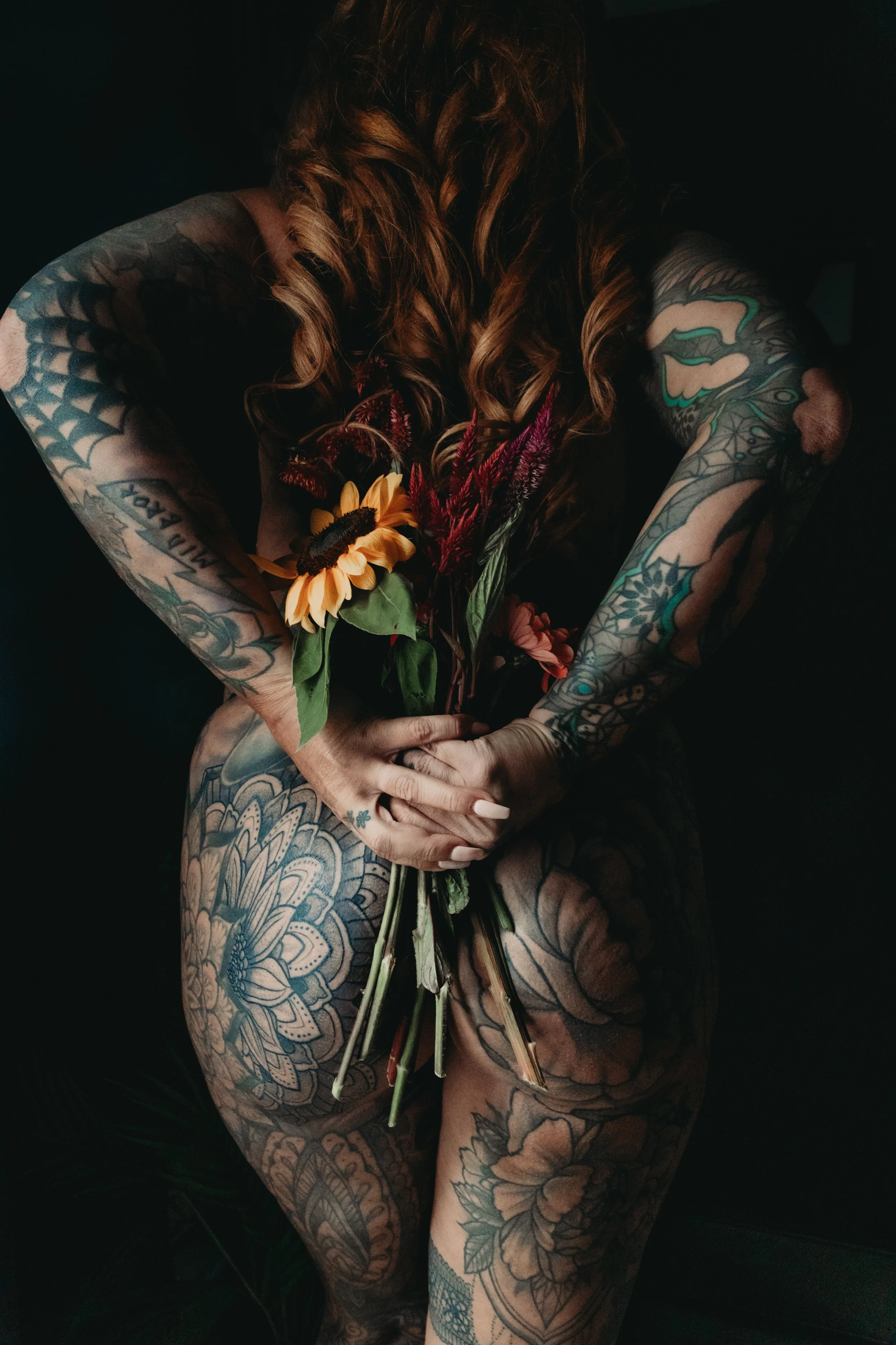 A person with long curly hair and extensive tattoos holding a bouquet of flowers over their lap, seated against a dark background.