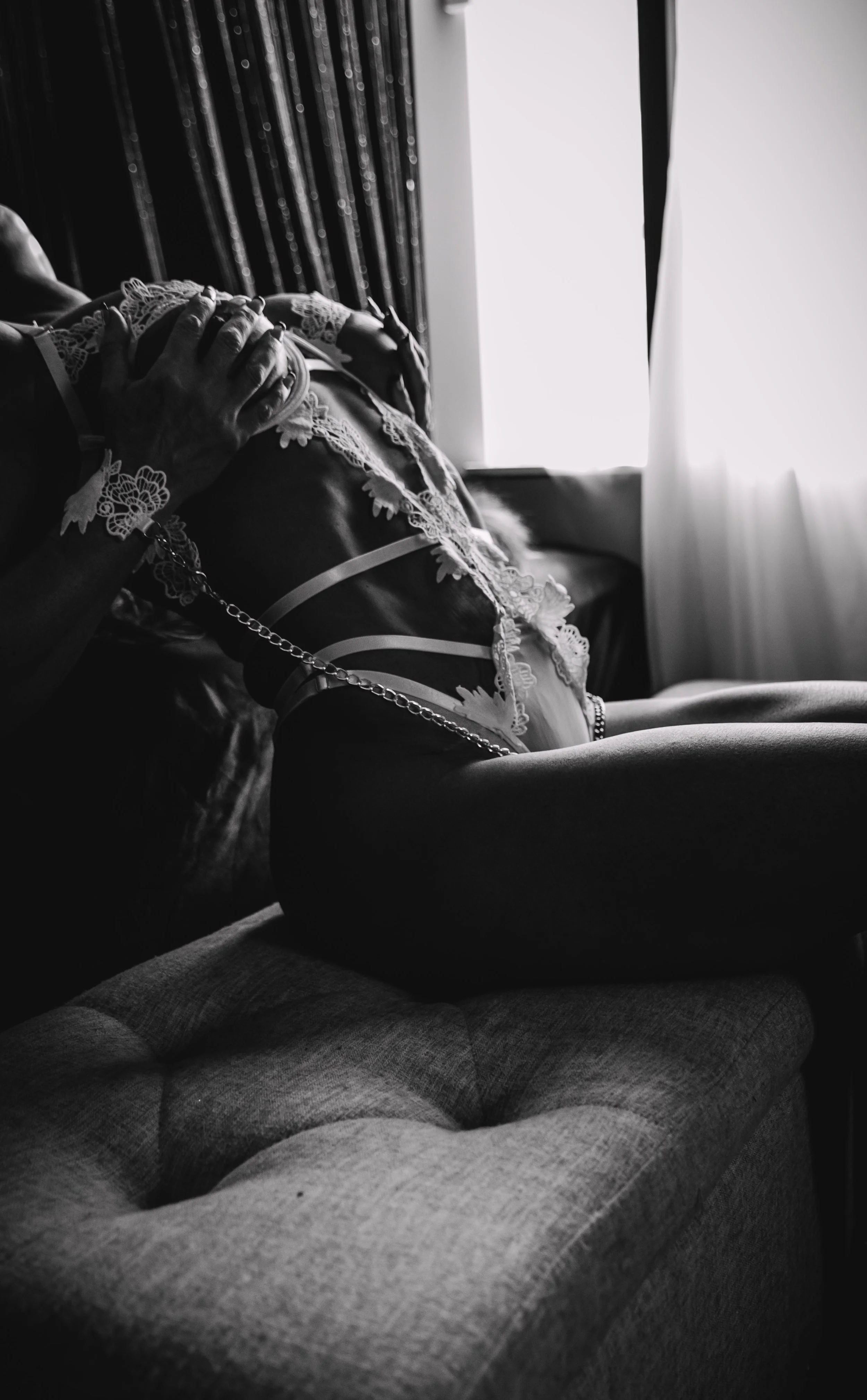 Black and white photo of a pregnant woman in lingerie sitting on a couch, with hands gently resting on her belly. Light comes through curtains in the background.