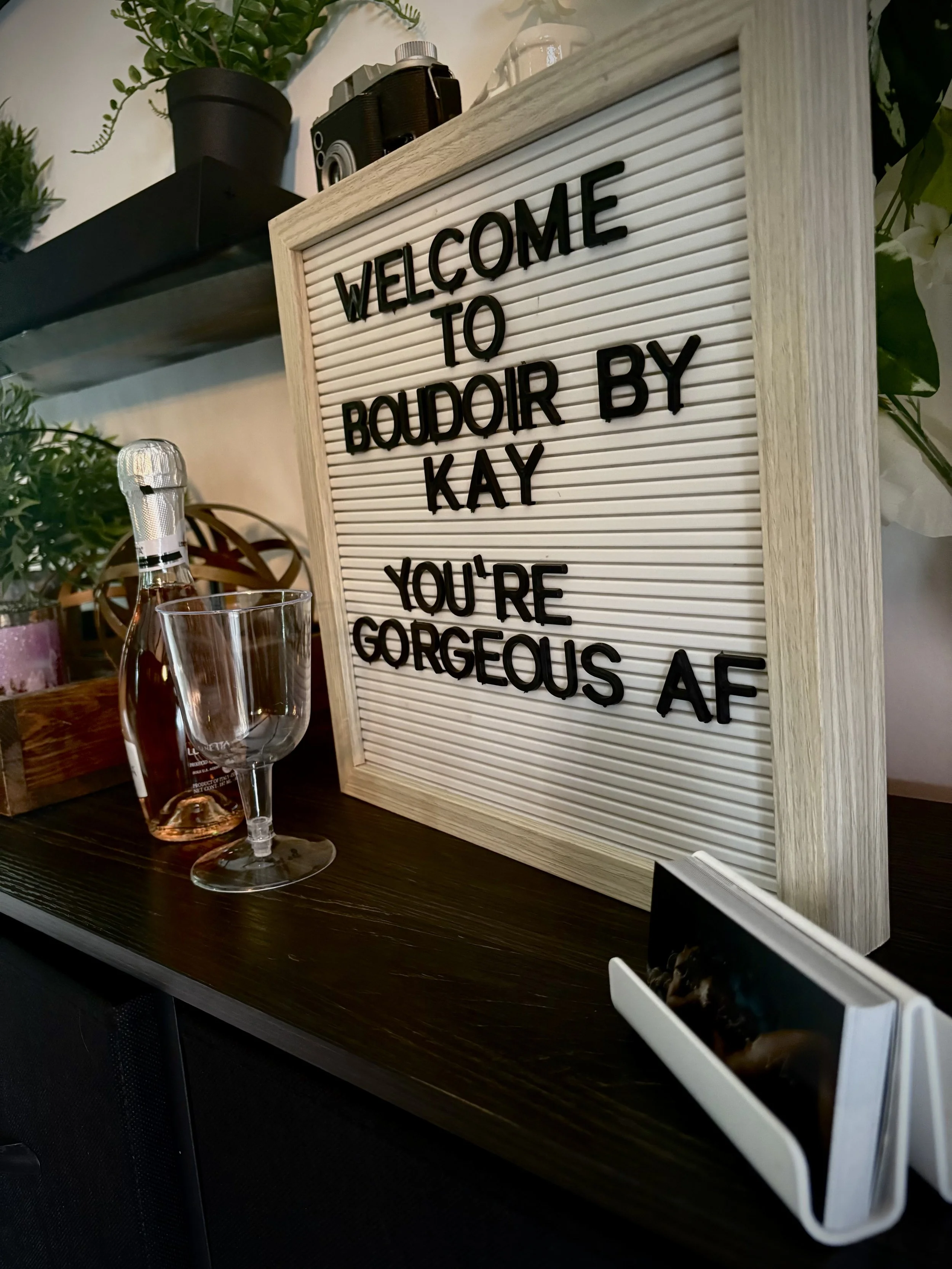 A wooden-framed letter board with black letters spelling out "Welcome to Boudior by Kay. You're gorgeous AF" on a dark wooden surface, next to a glass of rosé wine, a bottle of rosé, a small potted plant, a black camera, and a small photo in a white frame.