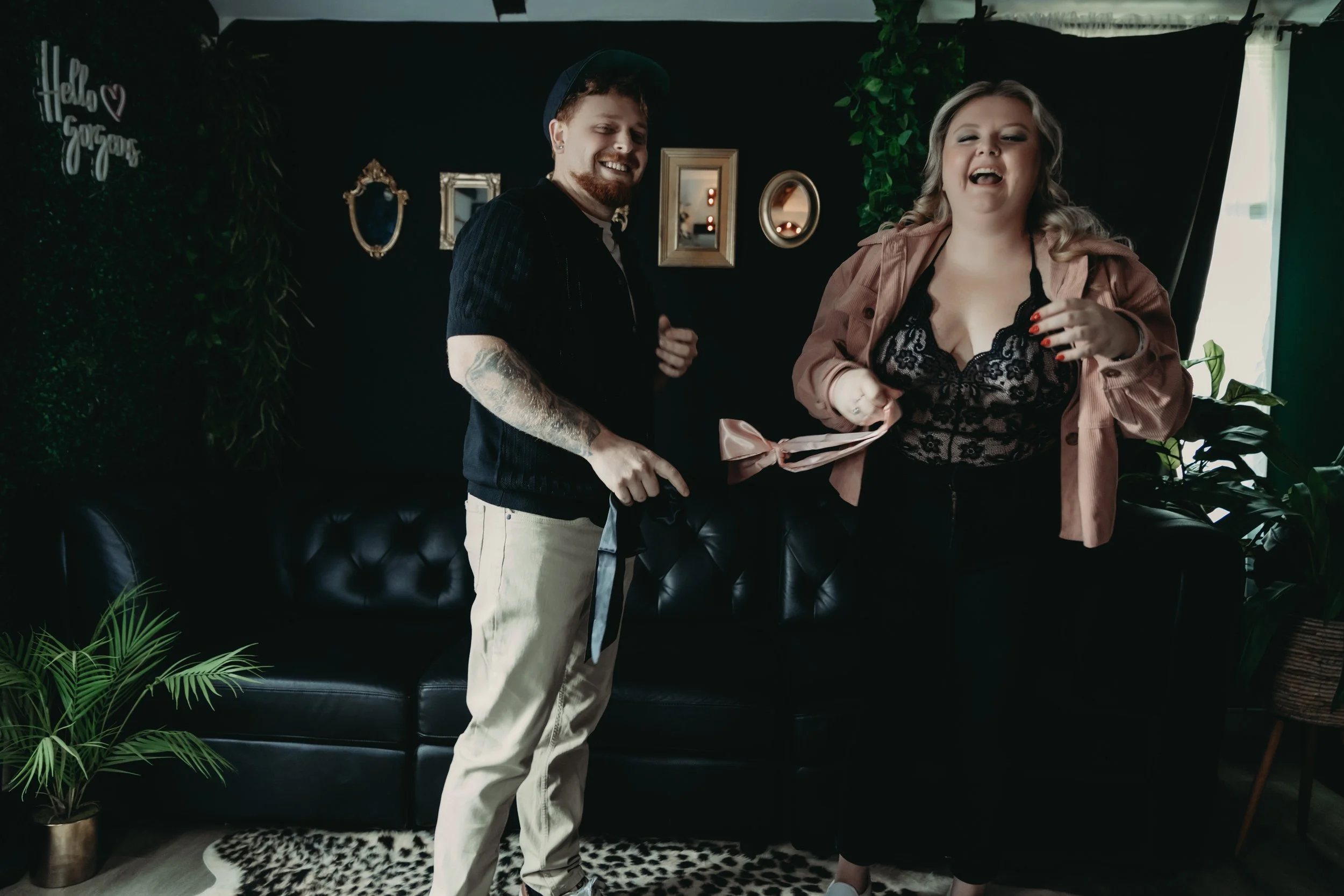 A man and woman with a baby standing in a living room, smiling. The woman is holding the baby, and the man is holding a small black item, possibly a gift or ribbon. The room has a black leather couch, green plants, and framed pictures on the dark wall.