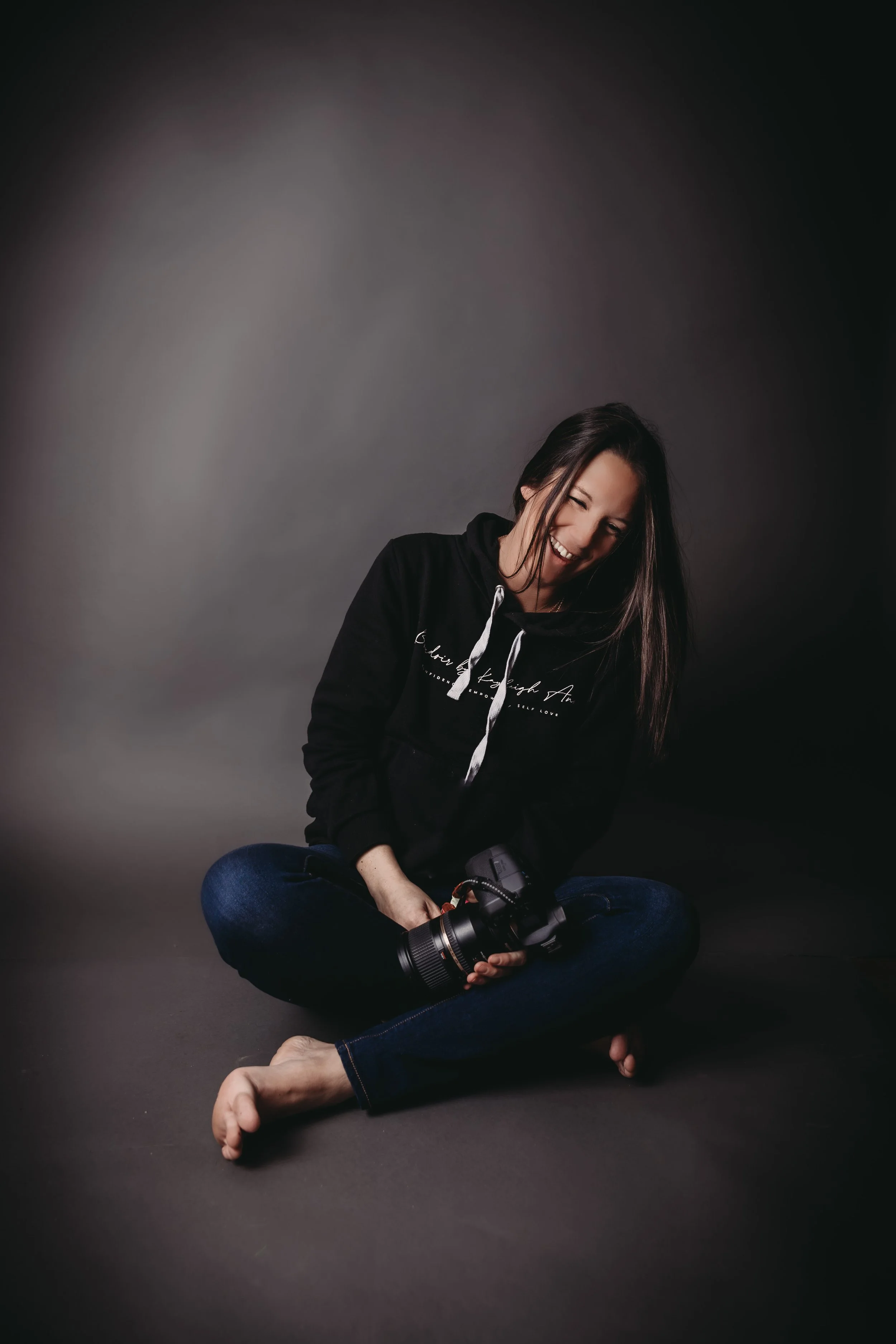 A woman sitting cross-legged on the floor, holding a camera with a large lens, smiling and looking down, wearing a black hoodie and blue jeans, with a dark studio background.