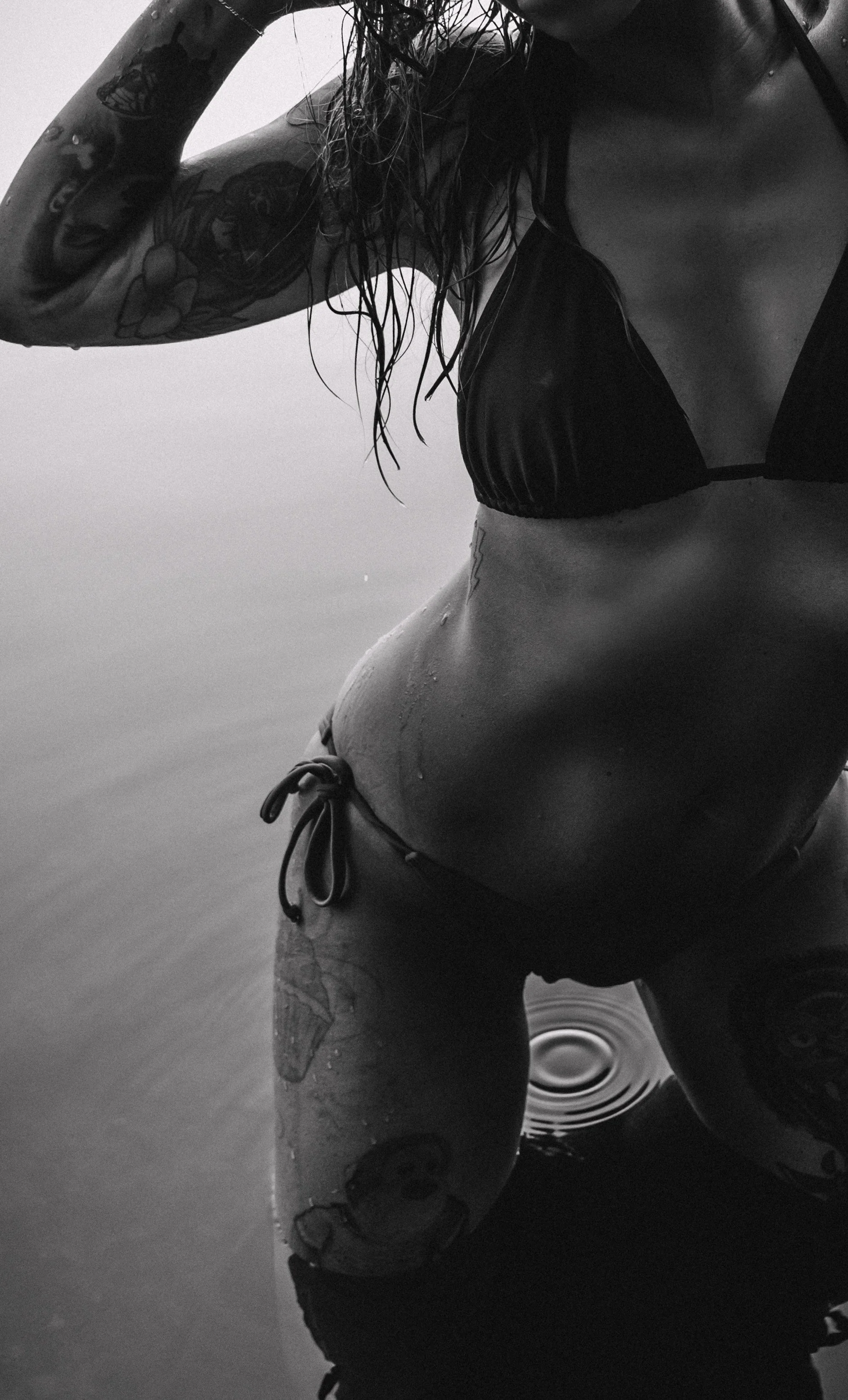 Black and white photo of a woman in a bikini top and bikini bottoms standing in water, with wet hair and tattoos on her arm and knee.