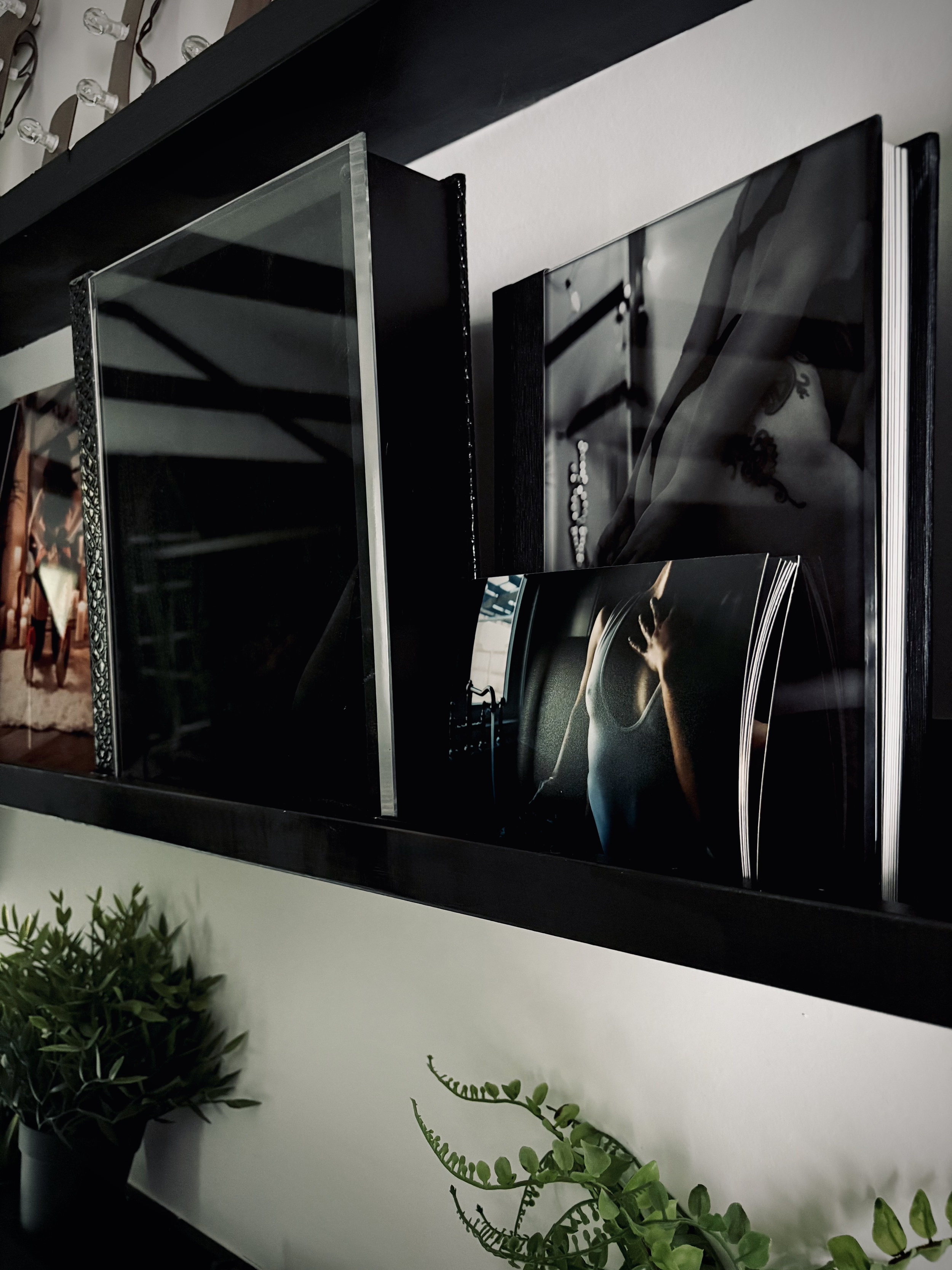 A black shelf holding black photo frames and books, with green potted plants at the bottom. The photo frames contain black and white photos, one showing a hand on a steering wheel.