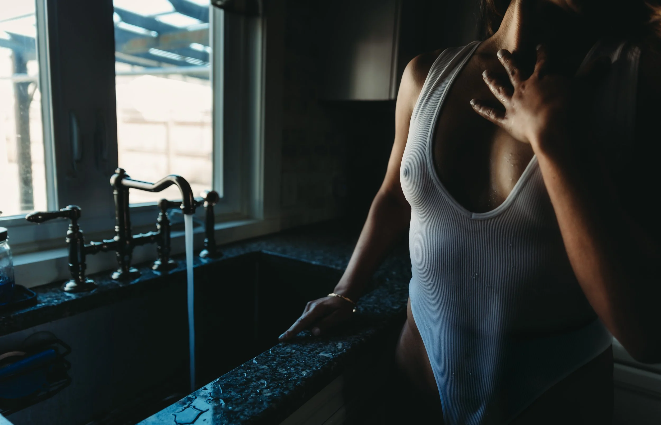 A woman in a white tank top is washing or rinsing her shirt in a kitchen sink, with sunlight coming through the window.