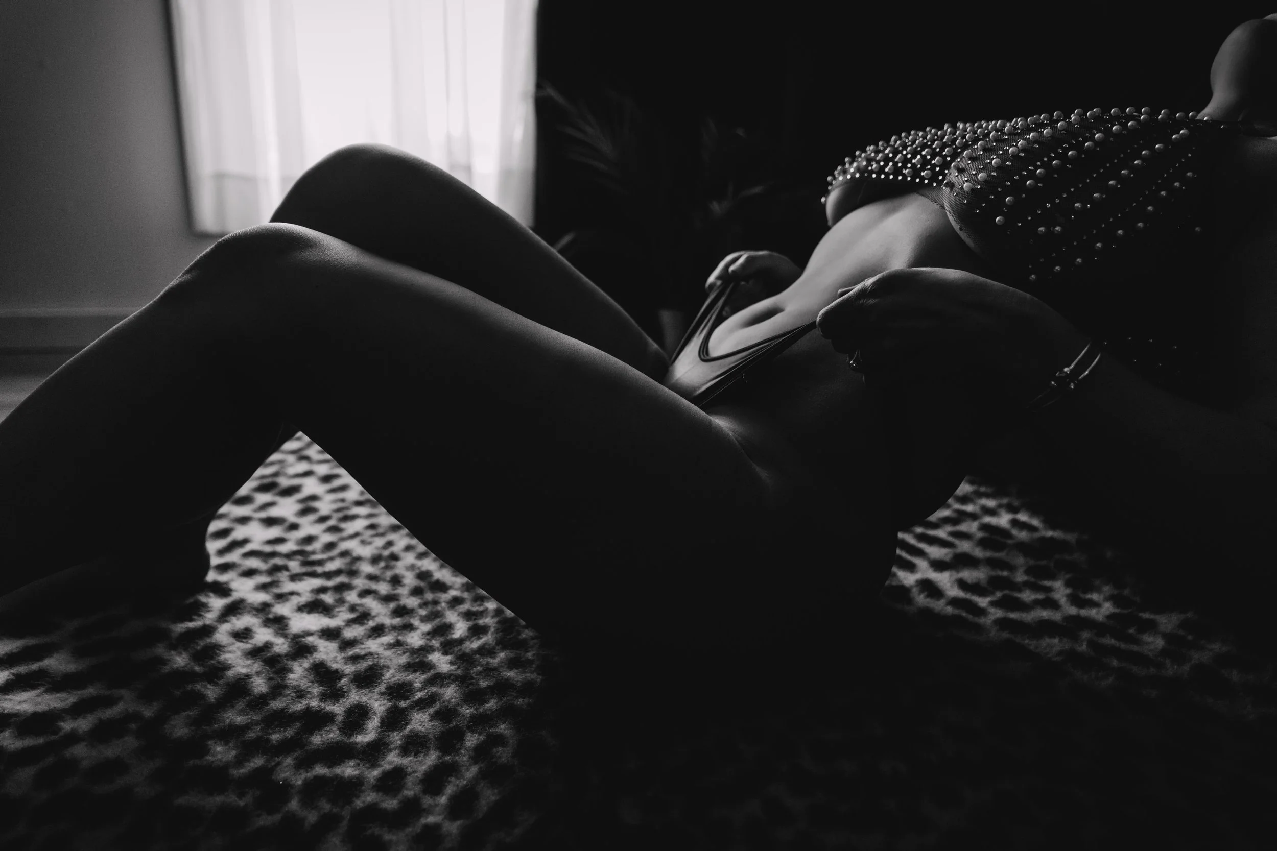 A person lying on a patterned carpet, wearing a studded top and fishnet stockings, in a dimly lit room with a window in the background.