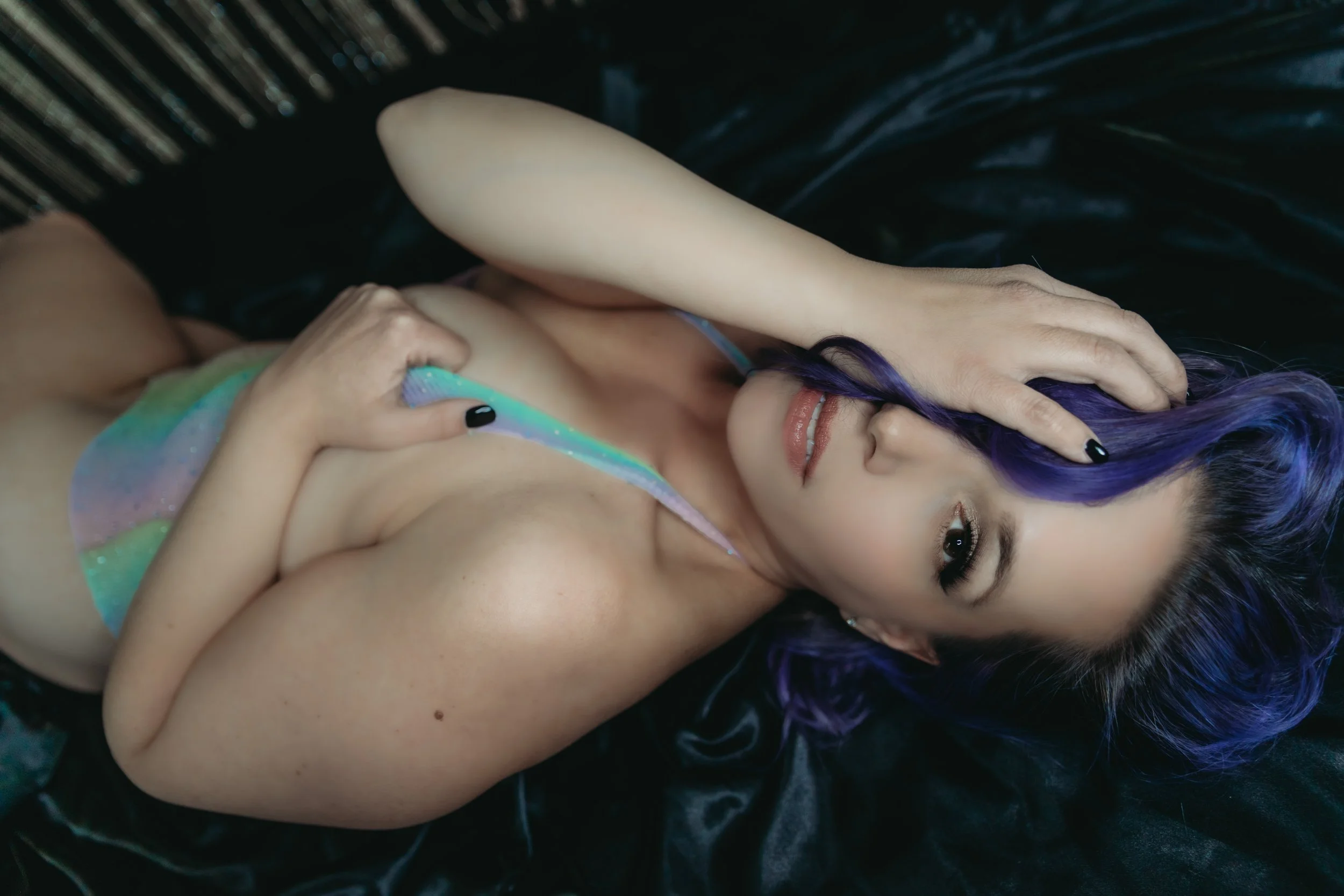 A woman with purple and black hair lying on black satin, covering her chest with one hand, wearing iridescent pasty on her body.
