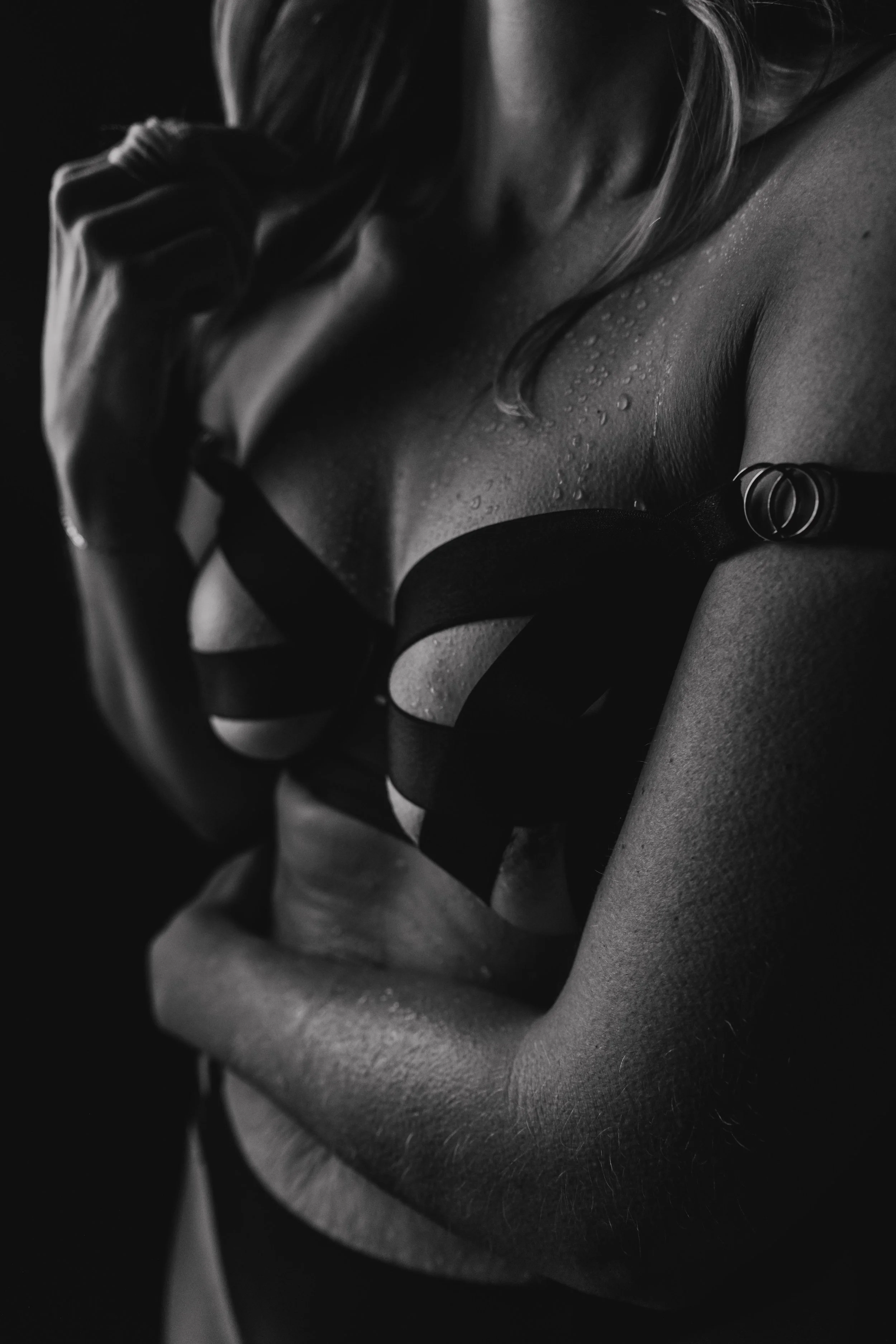 Close-up black and white photo of a woman wearing a strappy black lingerie top with water droplets on her skin, touching her face with one hand.