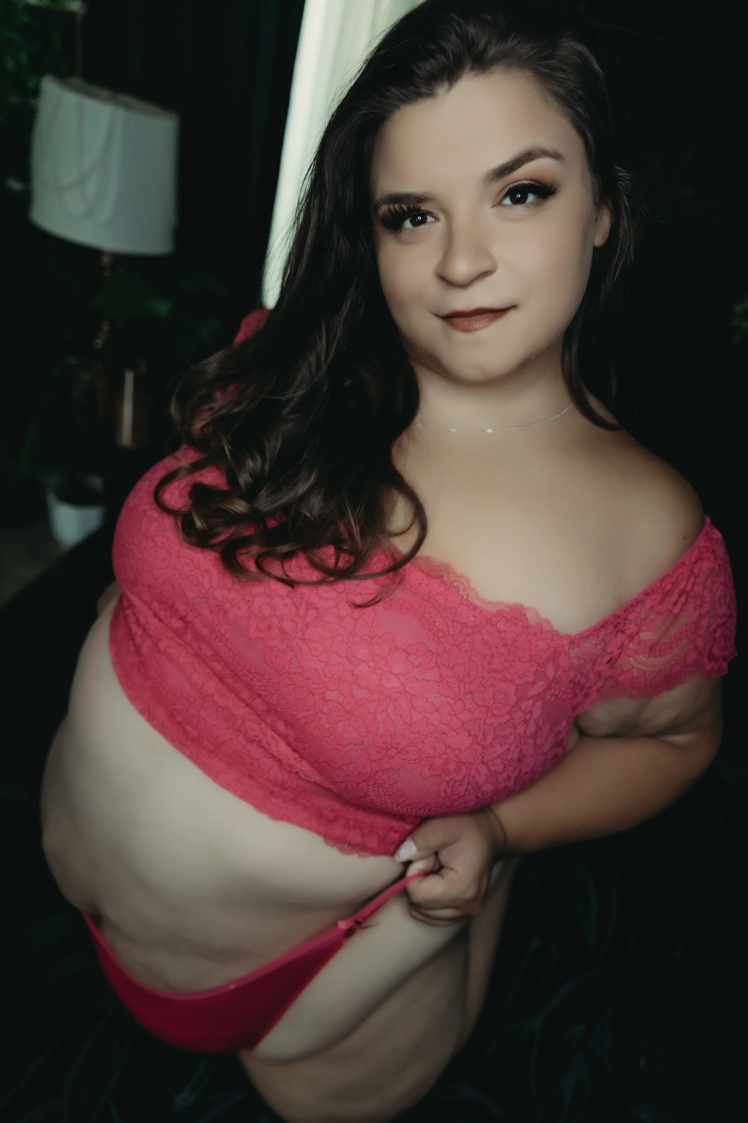 A woman with dark, curly hair and light skin wearing pink lace lingerie, sitting and holding her panties, with a dark background and a lamp and plant in the background.