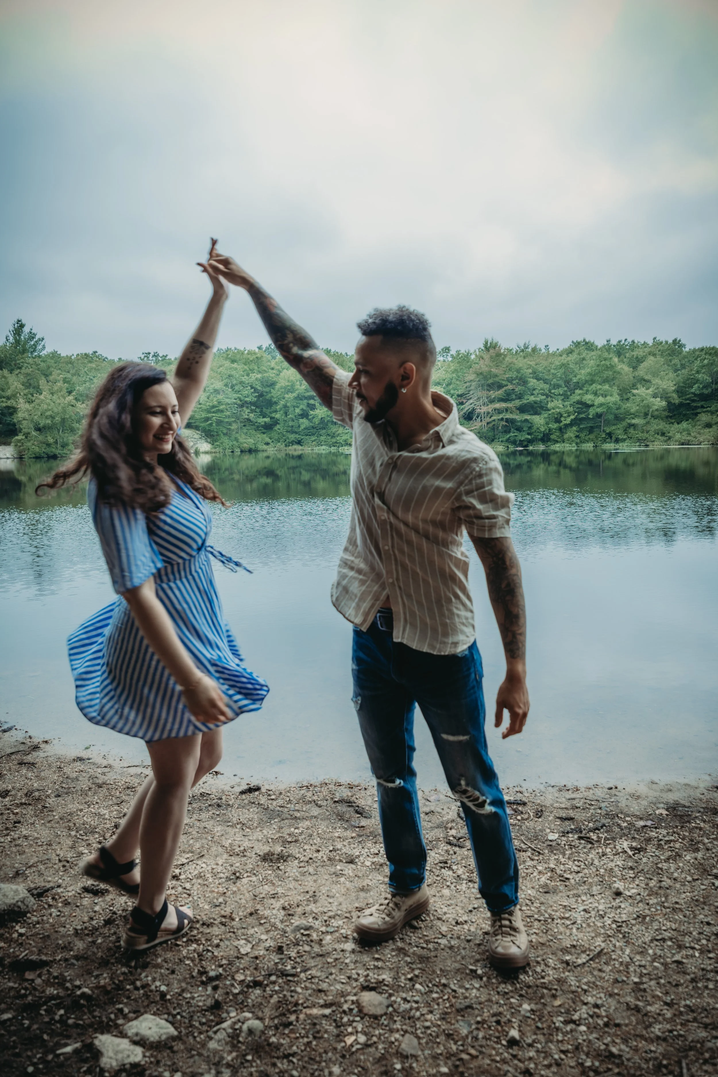 A couple dancing and smiling by a lake with trees in the background, holding hands up in a dance move.