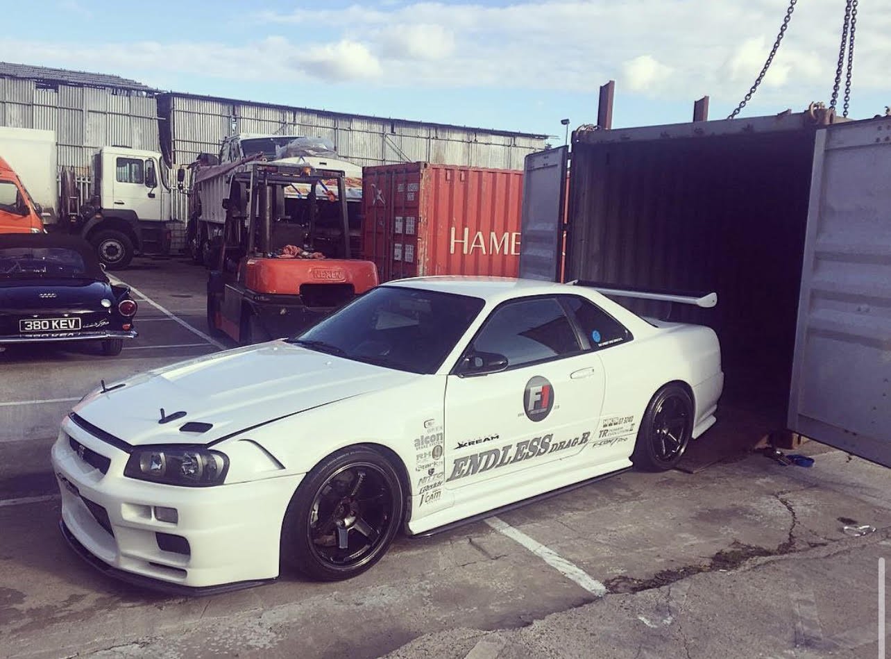 A white sports car with racing decals parked in a lot next to shipping containers, with other vehicles and a warehouse in the background.