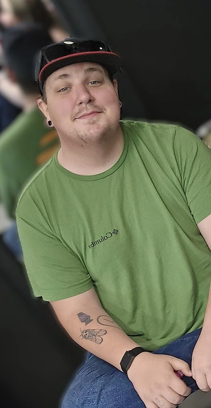 A young man with a tattoo on his left forearm, wearing a green t-shirt, a black cap, and a black wristband, smiling and sitting indoors.