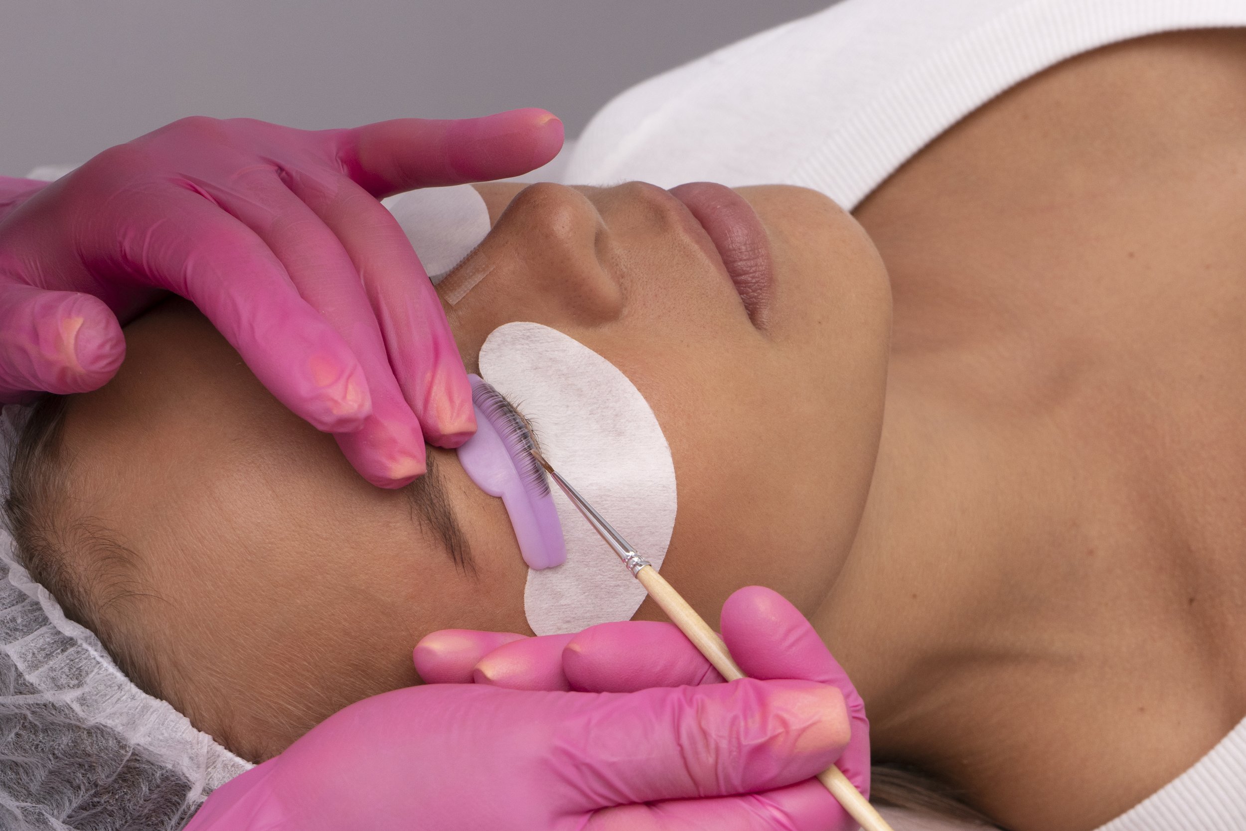 A person receiving a eyelash extension treatment, lying down with eyes closed, while a technician applies eyelash extensions with tweezers, wearing pink gloves.