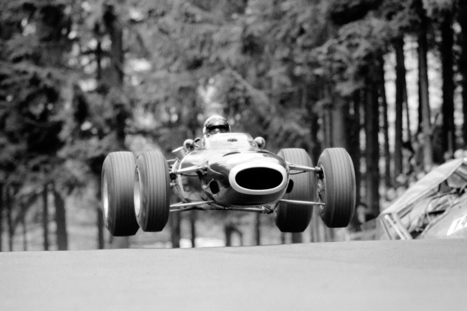 A black and white photograph of a vintage racing car in flight, seen from the front, with large tires and a streamlined body, surrounded by a forested background.
