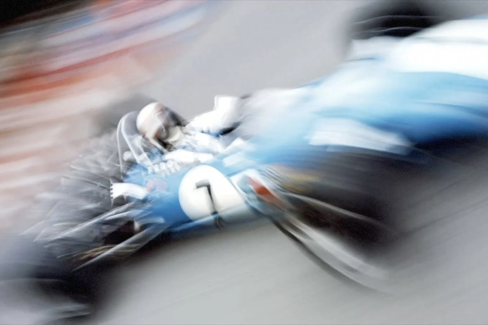 A vintage blue race car with the number 7 on it at high speed, with motion blur indicating fast movement.