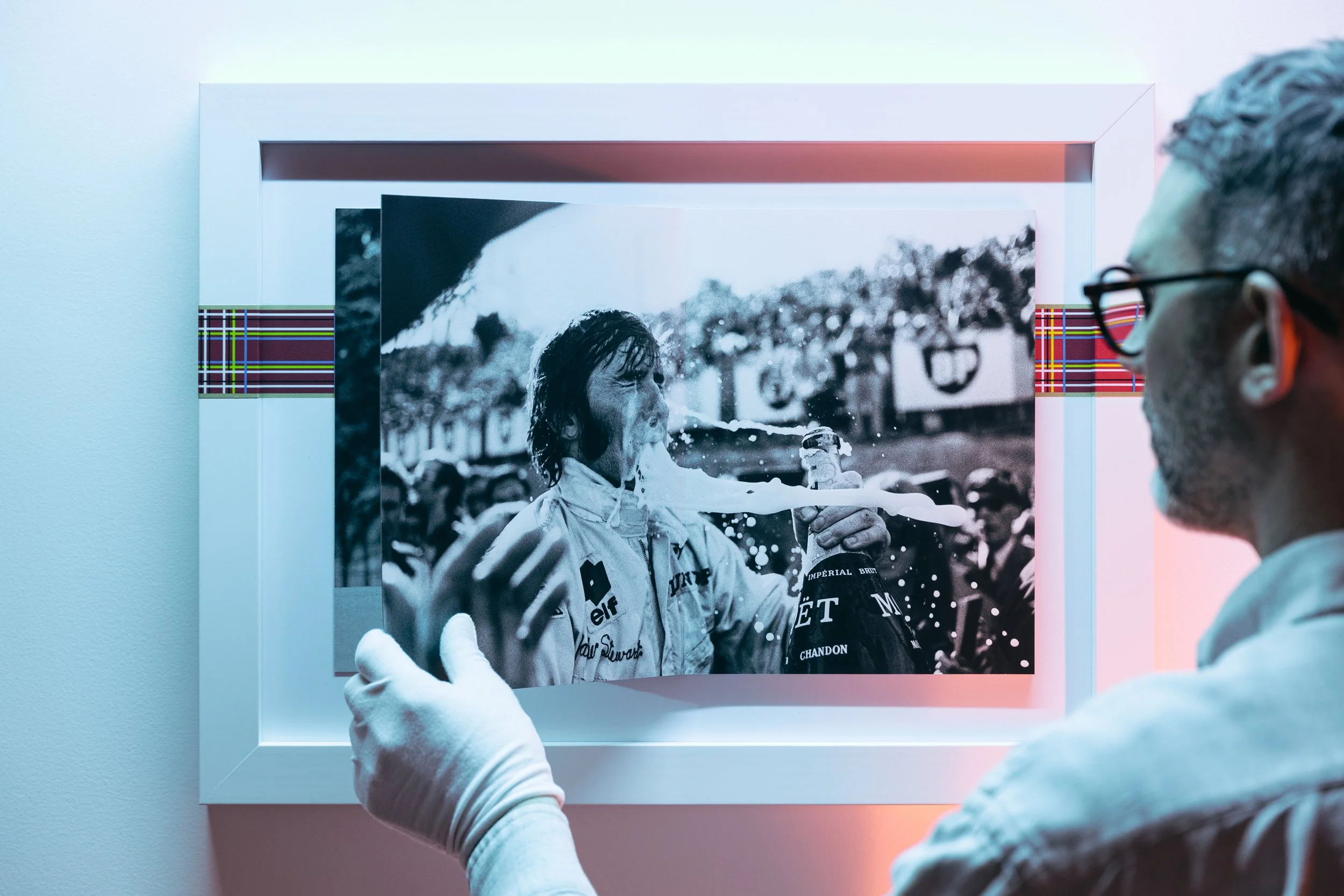 Person with gray hair and glasses holding a framed black-and-white photo of a woman spraying champagne, displayed on a wall.