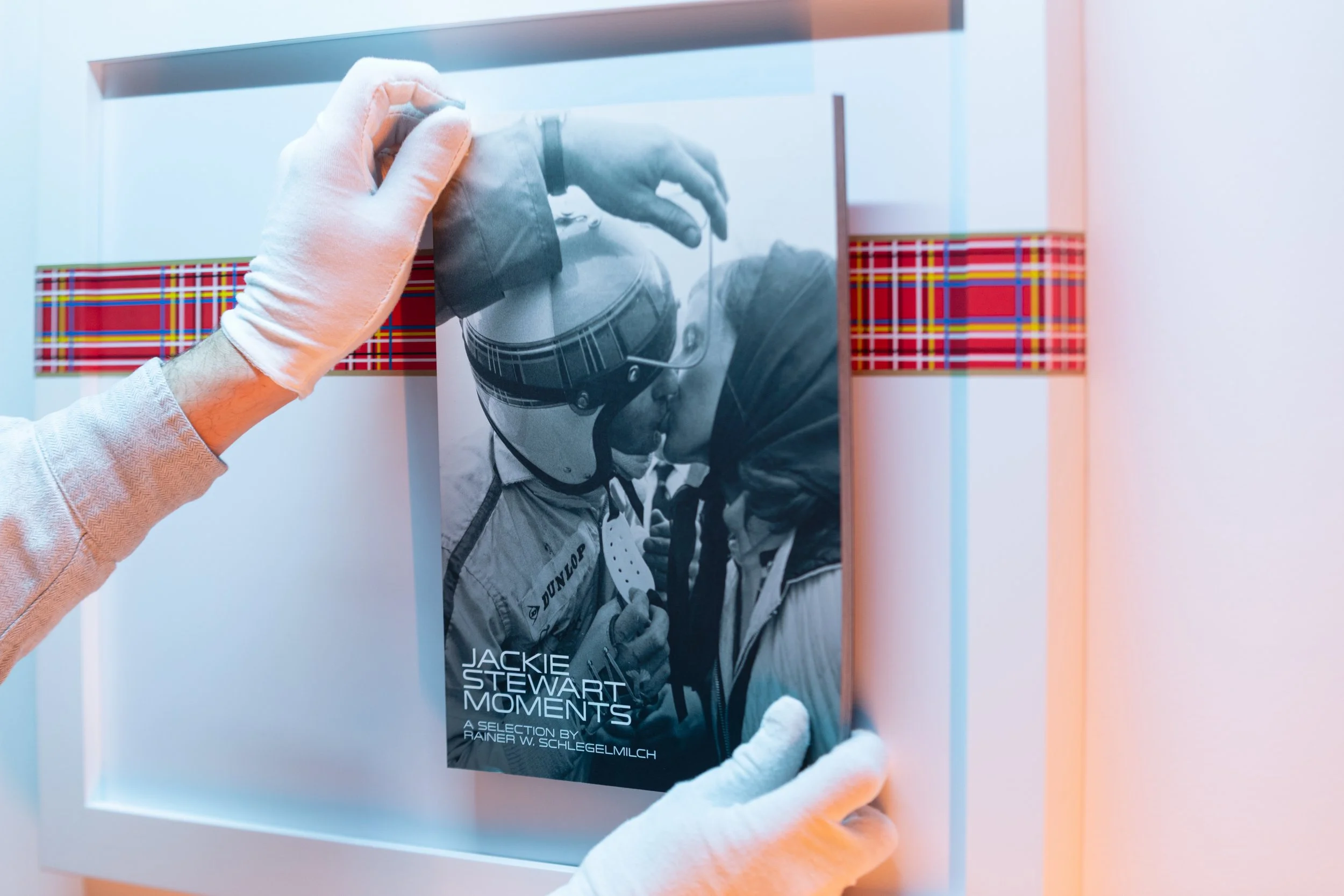 A person wearing white gloves holding a picture frame with a black and white photograph of a woman wearing a motorcycle helmet, and a book title "Jackie Stewart Moments".