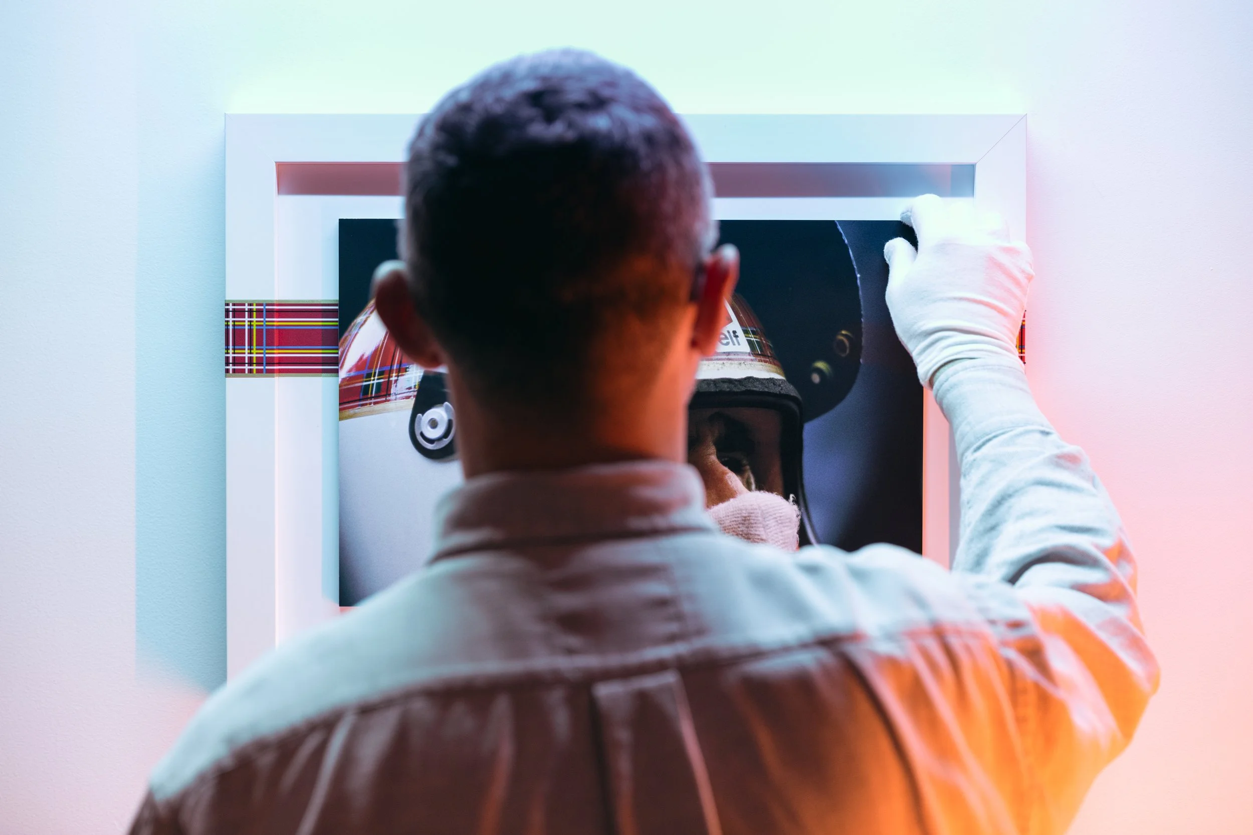 A person with short dark hair and wearing a plaid shirt is at an ATM, which has a patterned red, green, blue, and white design. The person is seen from behind, inserting a card or pressing buttons, with a white glove on their hand.