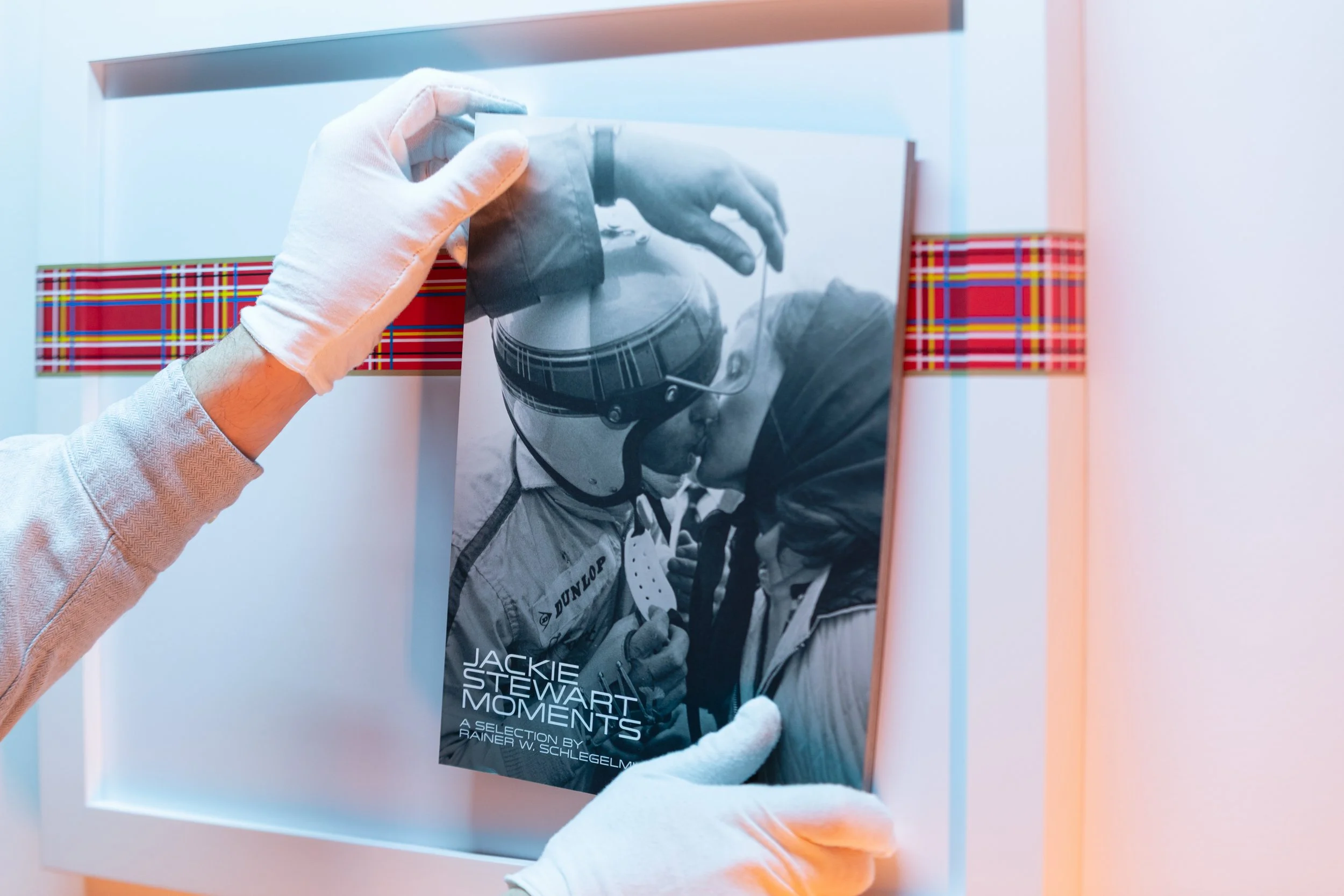 Close-up of a person's hands wearing white gloves, placing a black and white magazine titled "Jackie Stewart Moments" into a display case with a plaid border.