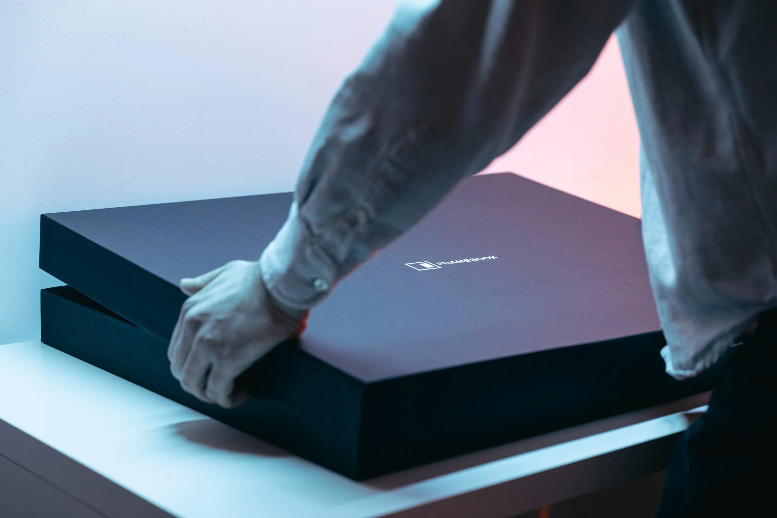 Close-up of a person in a gray leather jacket, reaching out with their right hand, resting on a black box labeled 'FRAMEBOOK' on a white surface, with pastel pink and blue lighting in the background.