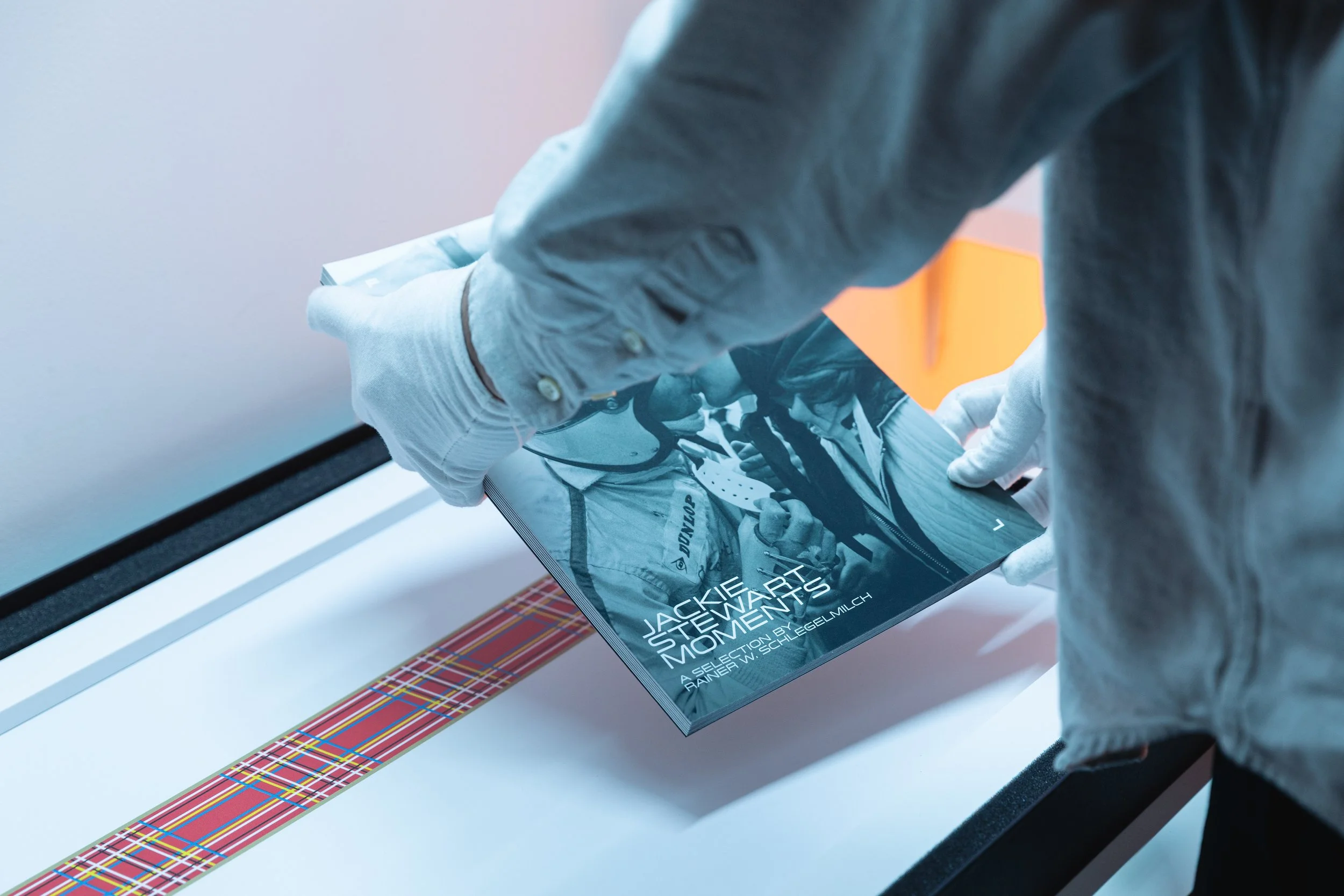 Person wearing gloves holding a book titled "Jackie Stewart Moments" with a photograph of Jackie Stewart on the cover. The book is placed on a surface with a tartan-patterned border.
